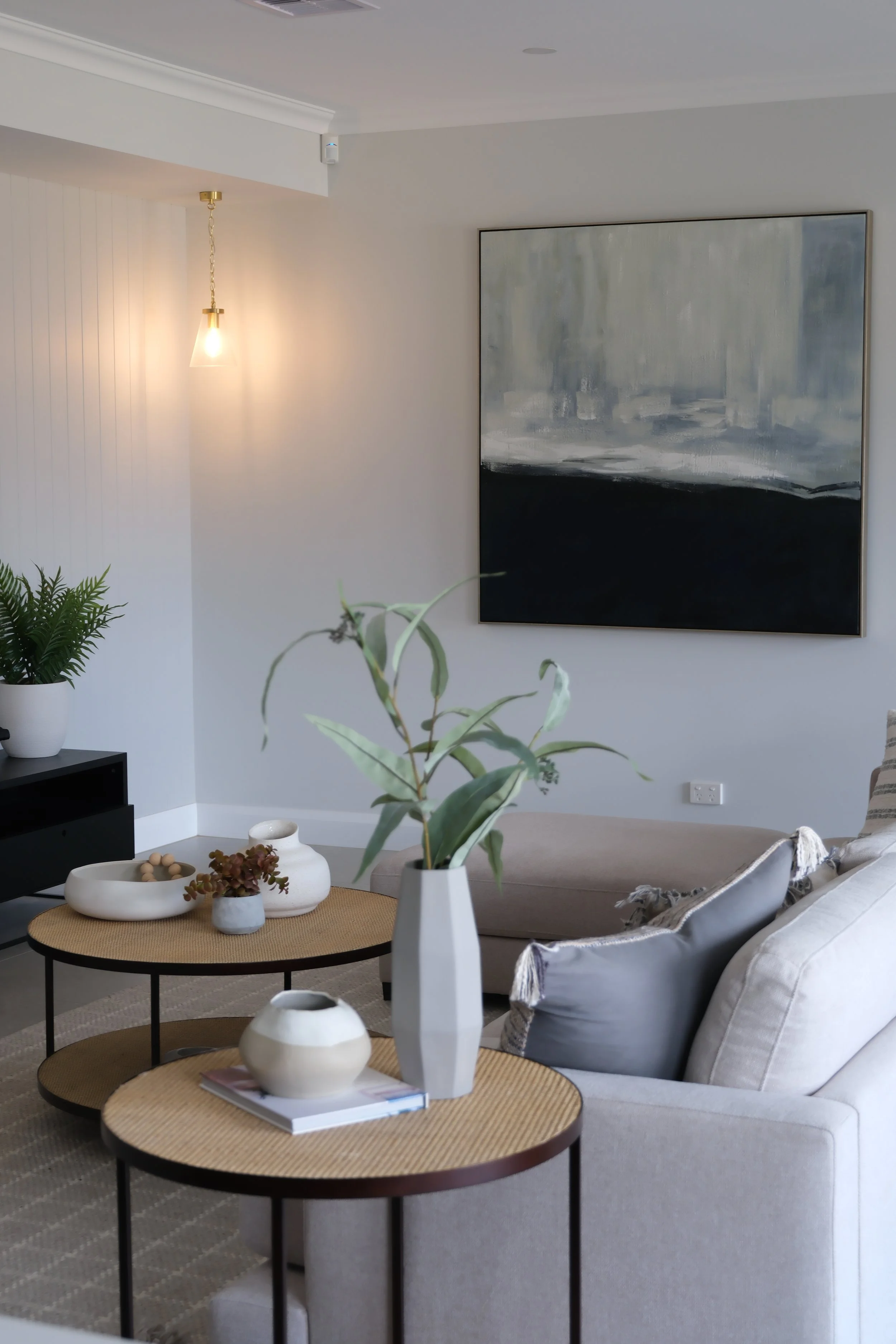 A modern living room with a light grey couch, a round coffee table with beige woven top, and decorative vases on it. There is a large abstract painting with shades of grey and black on the wall. A potted plant is on the left, and a hanging light fixture with a glass shade is on the ceiling. The room has a neutral color scheme and contemporary decor.
