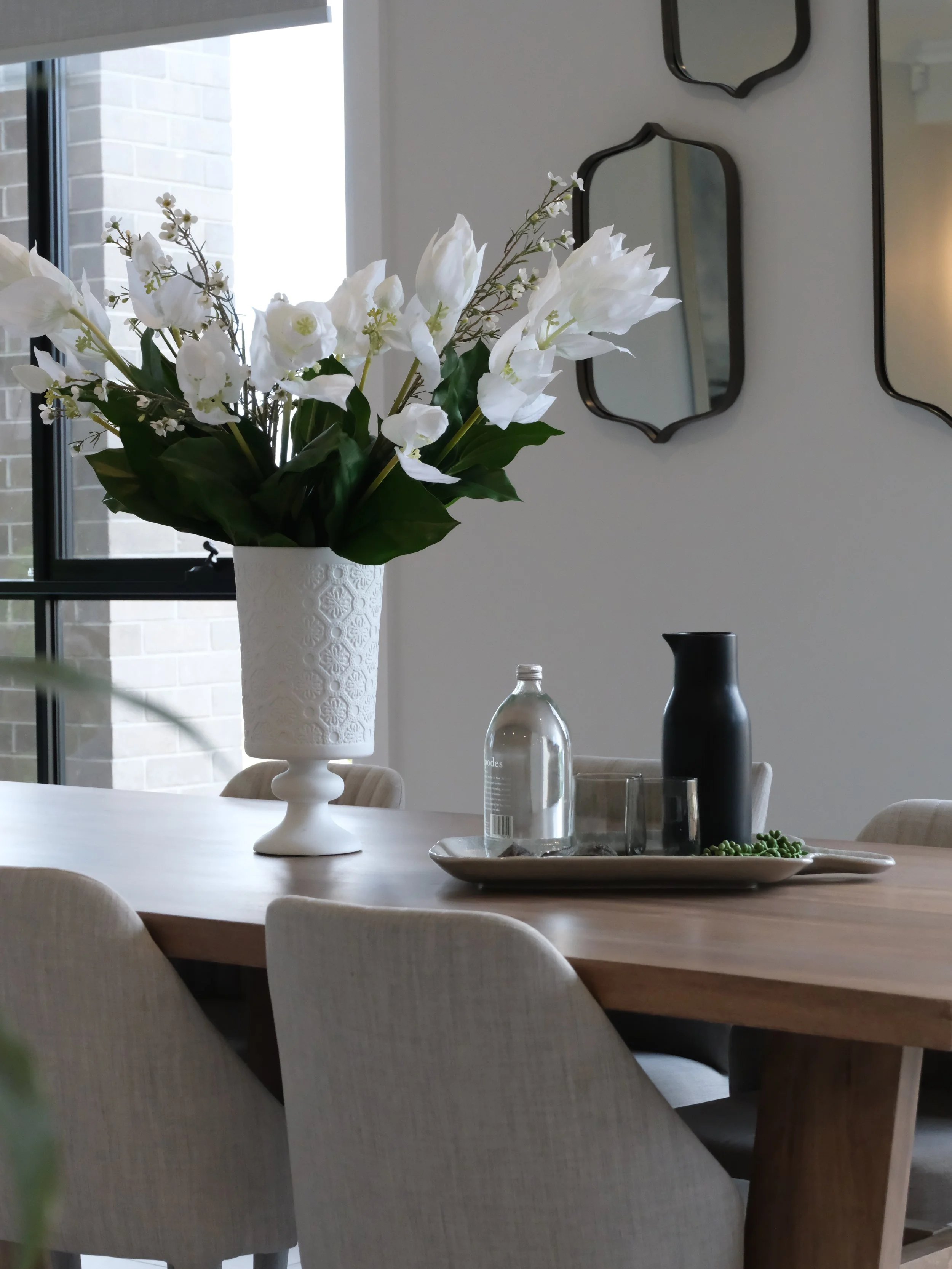 A dining room table with a white vase holding white flowers and greenery, along with a glass water bottle, a black vase, and a tray with decorative items. There are large windows and three black-framed mirrors on the wall.