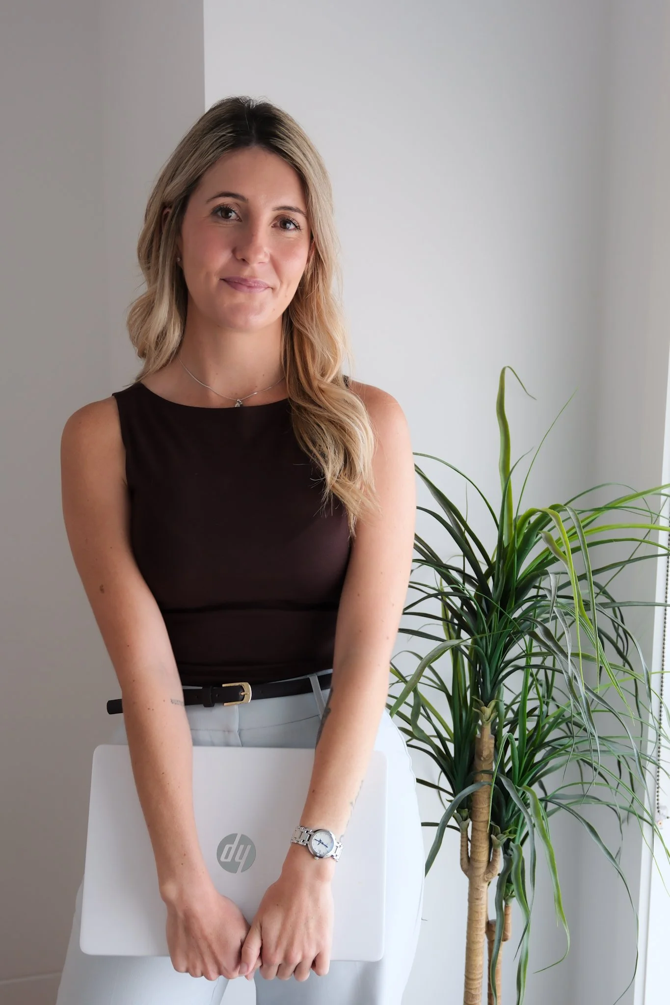 A woman with blonde hair wearing a black sleeveless top and white pants standing behind a white HP laptop, with a large green potted plant next to her in a modern white-walled room.