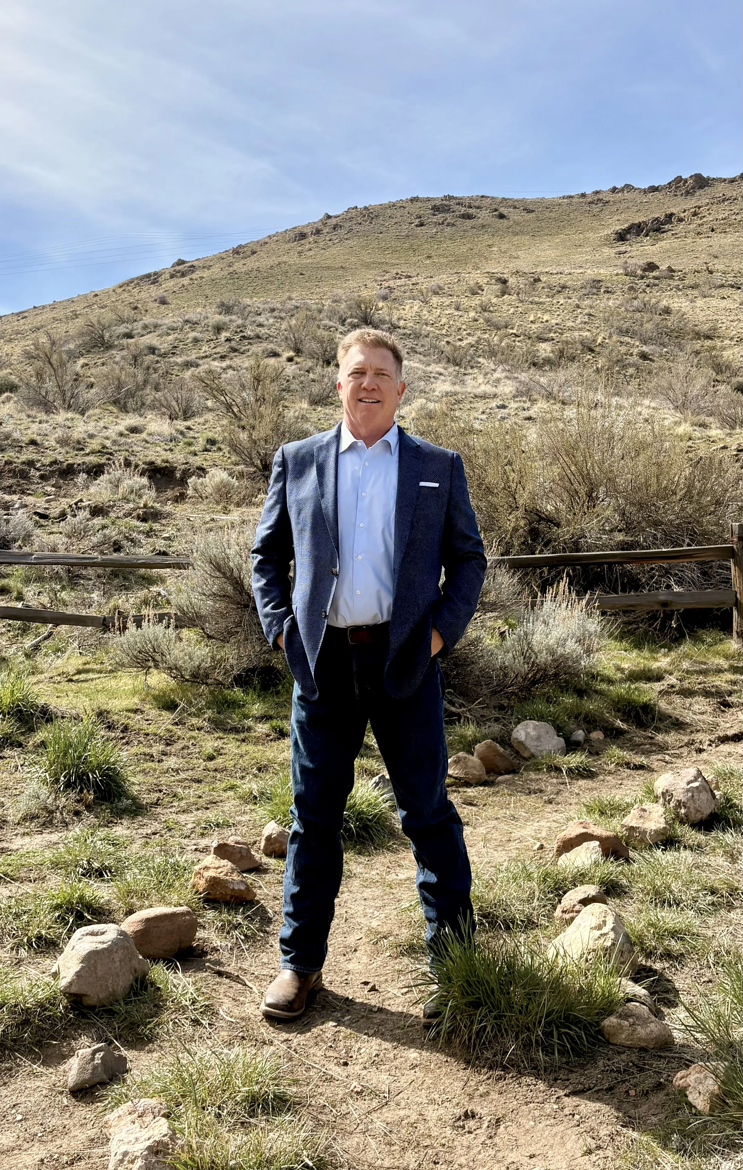 A man in a blue suit jacket, white shirt, and jeans stands outdoors in a desert landscape with dry bushes, rocks, and a hill in the background, under a clear blue sky.