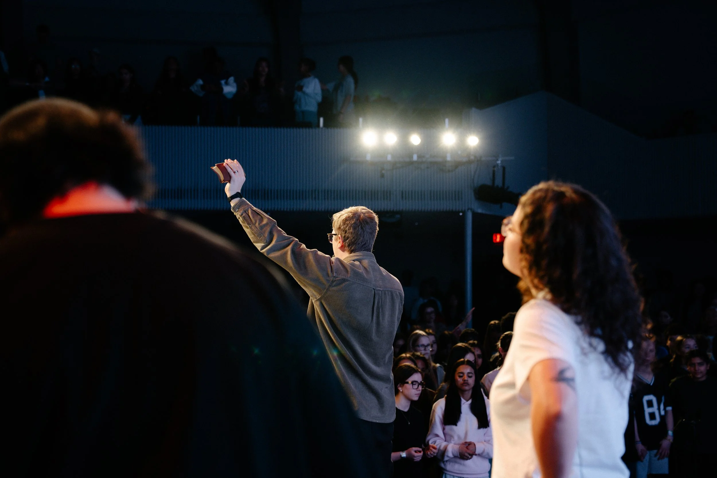 Audience members watching a performance or presentation in a dark auditorium, with a man holding up a book or paper in the foreground and stage lights illuminating the scene.