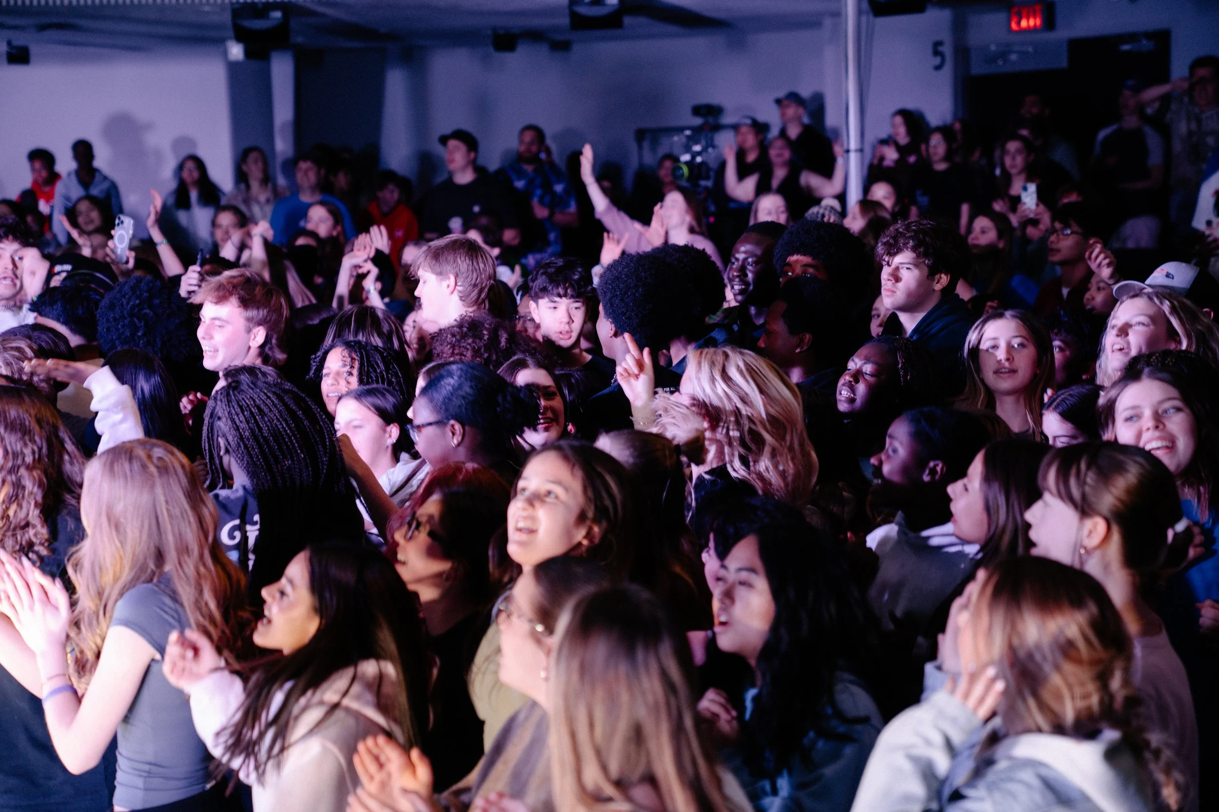 Crowd of young people at an indoor event, some raising their hands, with a camera on a tripod and an exit sign in the background.