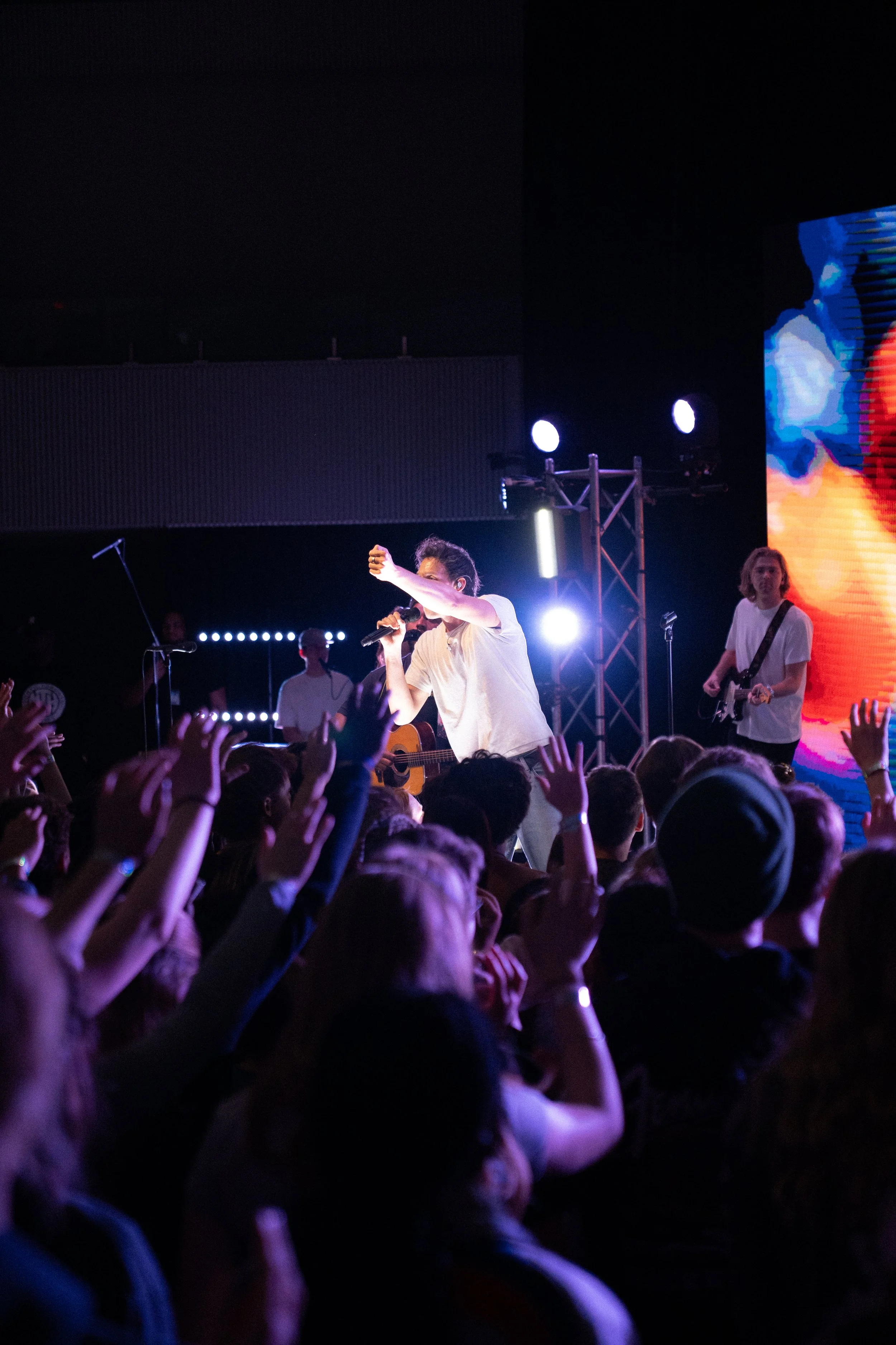 A crowd of people at a concert or live performance, with a singer and guitarist on stage under bright lights and a large screen display showing colorful visuals.