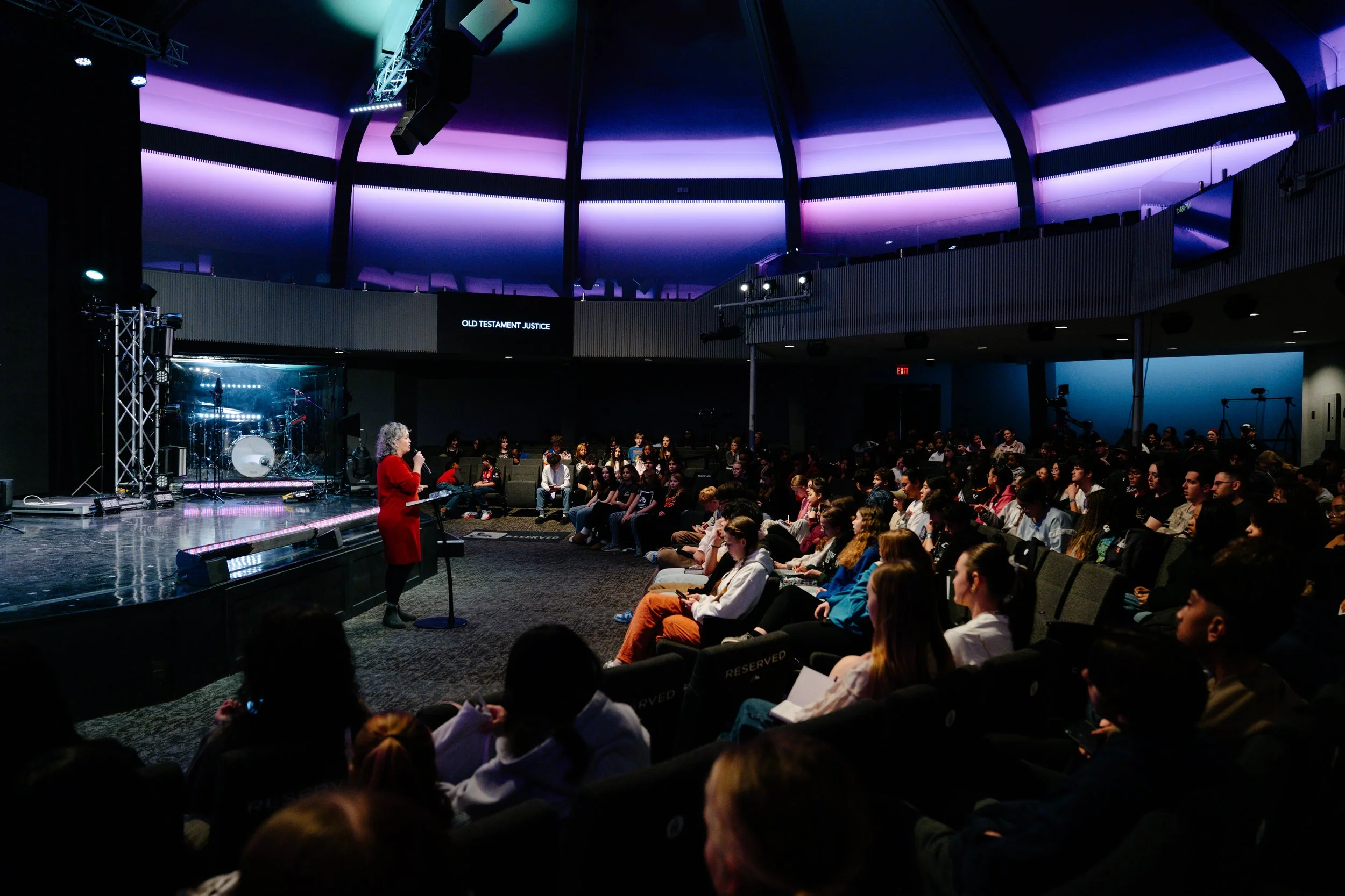 A woman in a red dress speaking into a microphone on stage in front of an audience inside a modern auditorium with purple and blue lighting. A screen behind her displays the words 'Old Testament Justice.'