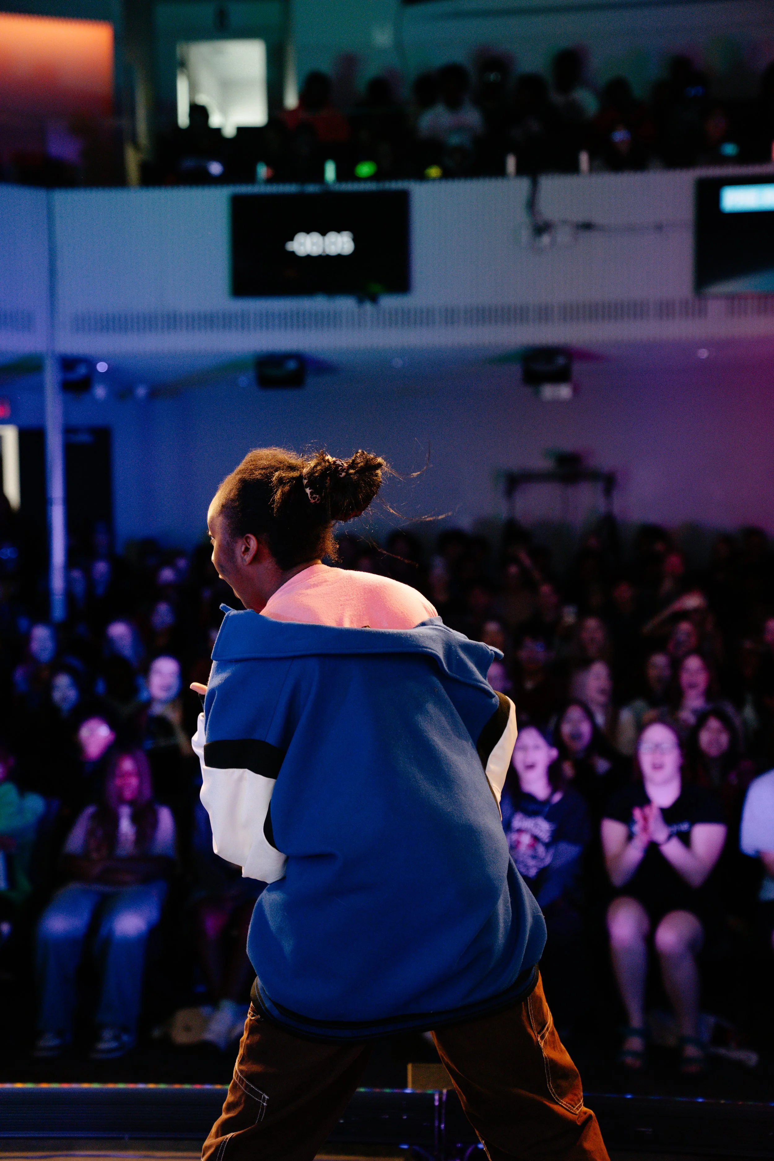 A person with curly hair tied up in a bun wearing a blue jacket and dark pants stands on stage in front of a large audience. The audience is seated and visibly engaged, with many appearing to smile or laugh. The background shows multiple screens and 