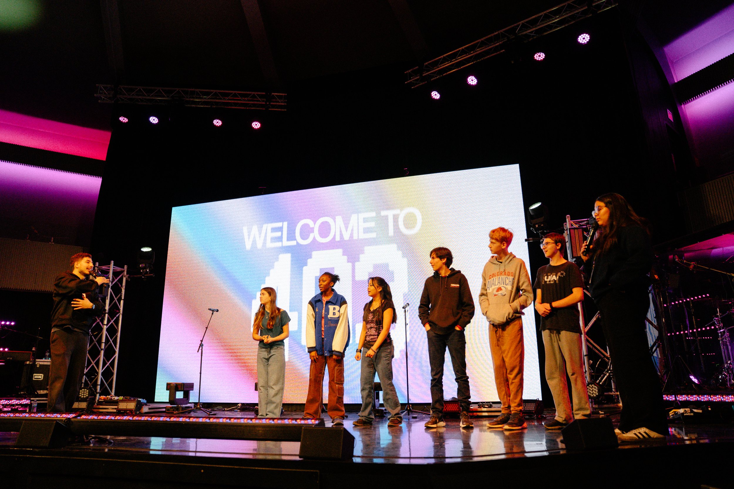 A group of young people standing on a stage during a presentation, with a woman holding a microphone to the right, and a large screen behind them that says 'Welcome to' with a colorful background.
