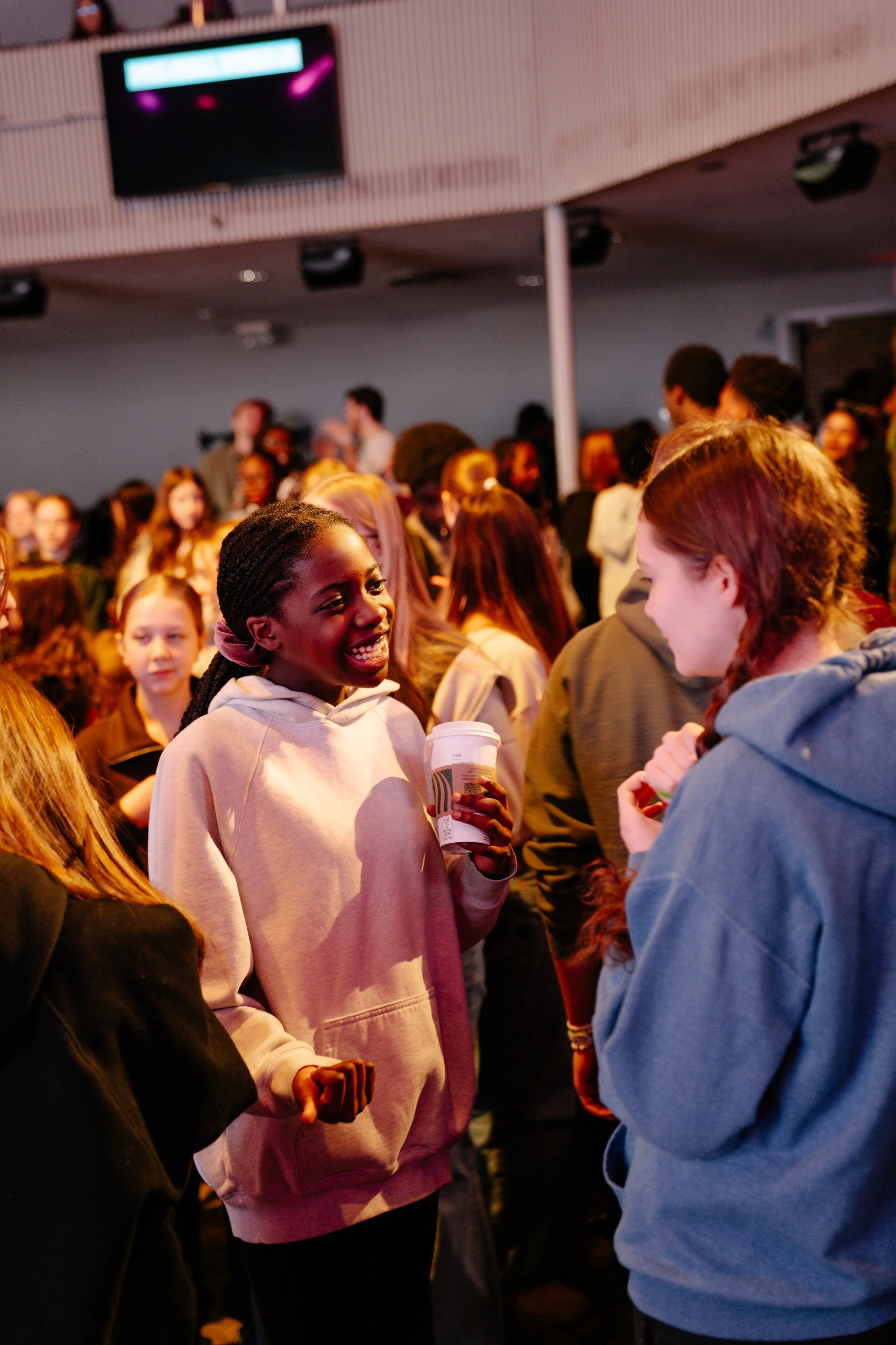 A group of people gathered at an indoor event. In the foreground, a woman with braids and a light-colored hoodie is smiling and holding a coffee cup. Others are engaged in conversation, with a crowd in the background and a large screen on the wall.