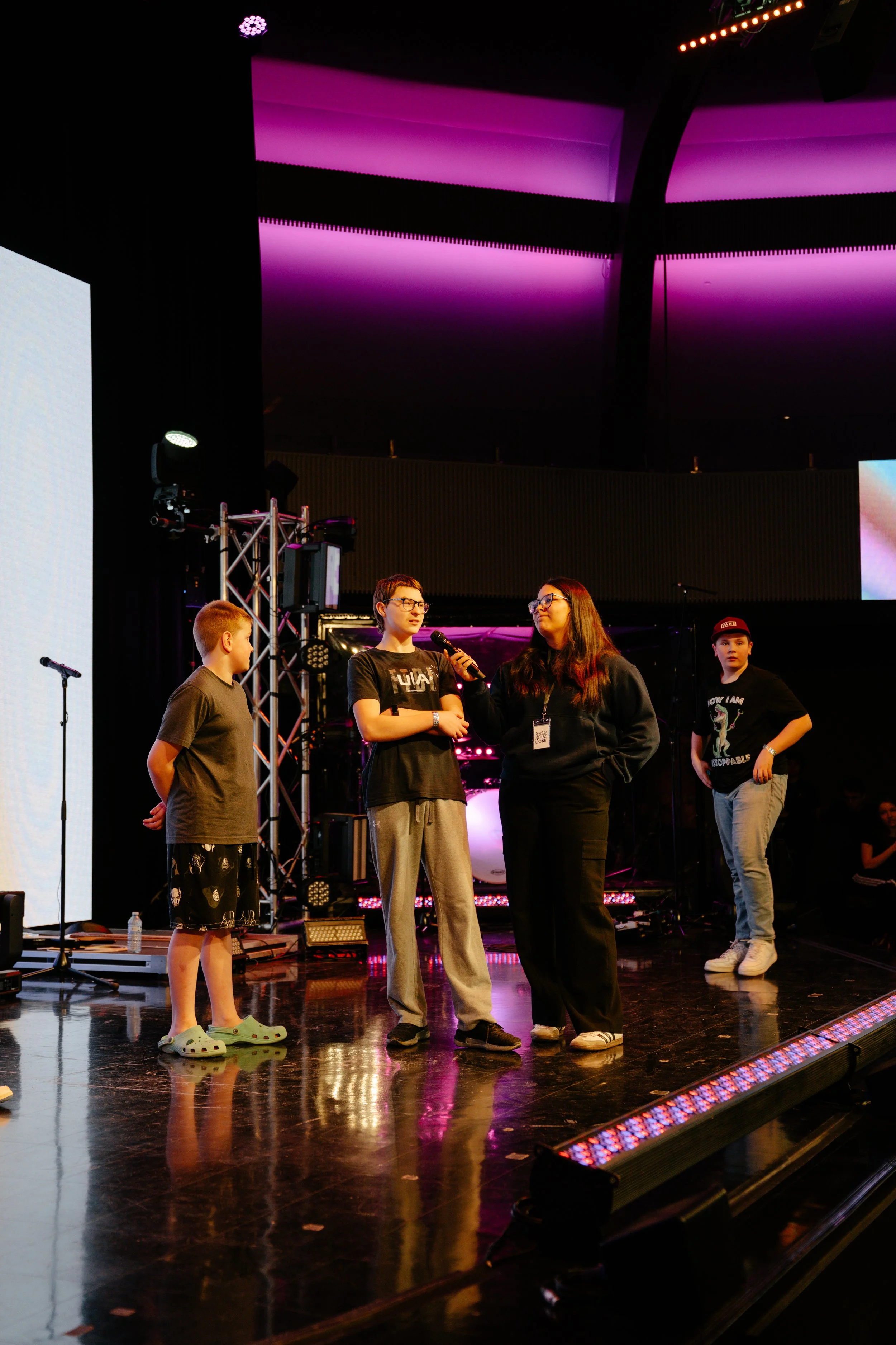 A group of five young people standing on a stage with colorful lighting and large screens behind them. One person is speaking into a microphone, and others are standing nearby, some looking at the speaker. The stage has equipment and a reflective flo