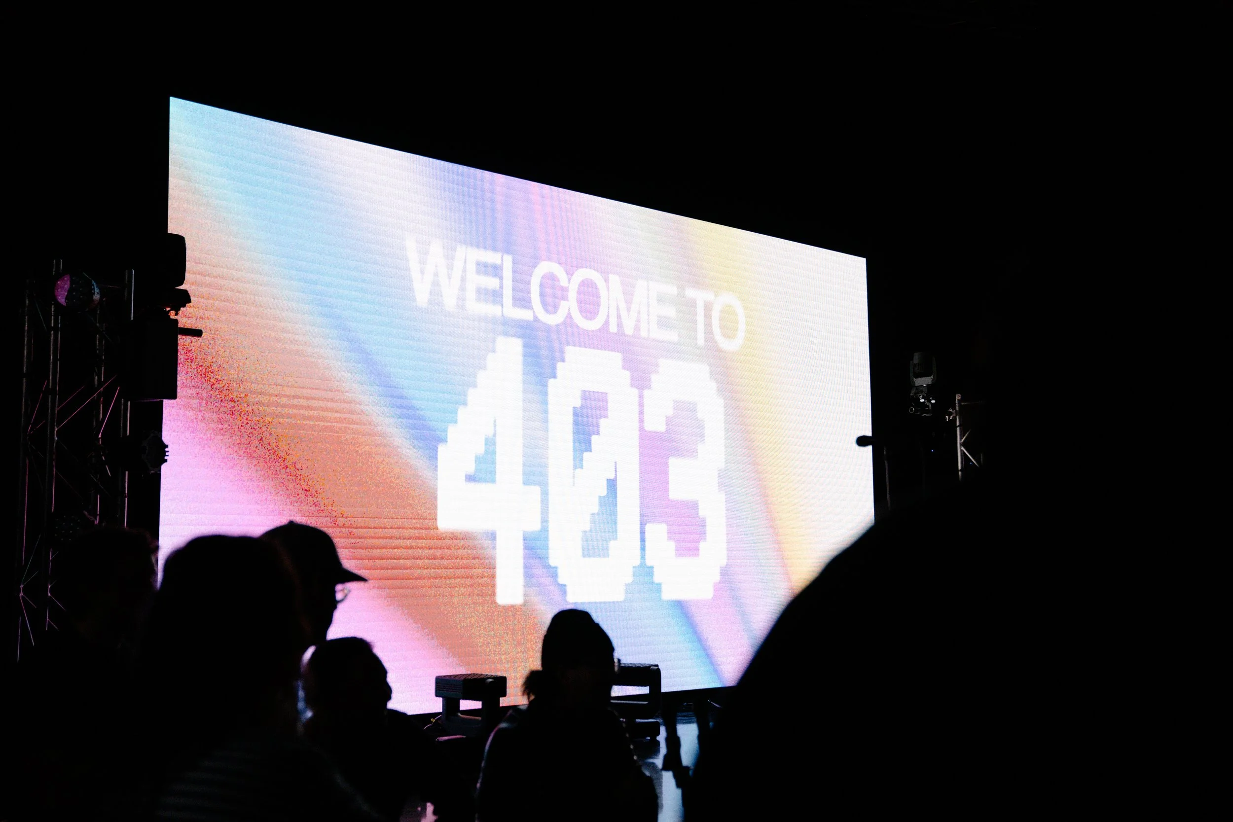 Large illuminated screen displaying the words 'Welcome to AOE' with pixel art graphics, in a dark setting with silhouettes of people in the foreground.