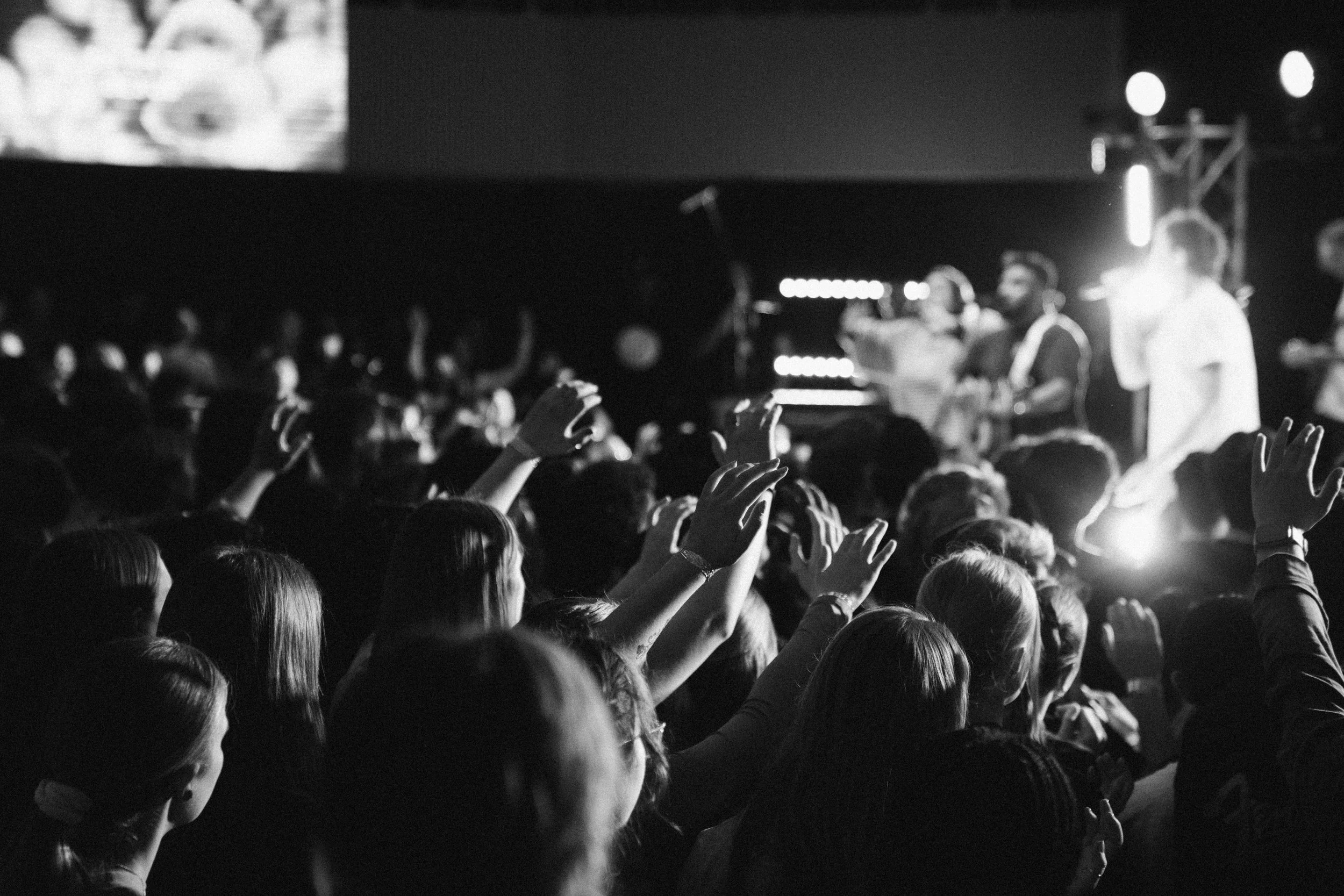 Black and white photo of a concert or event with a crowd of people raising their hands and facing a stage with performers or speakers, bright lights are visible in the background.