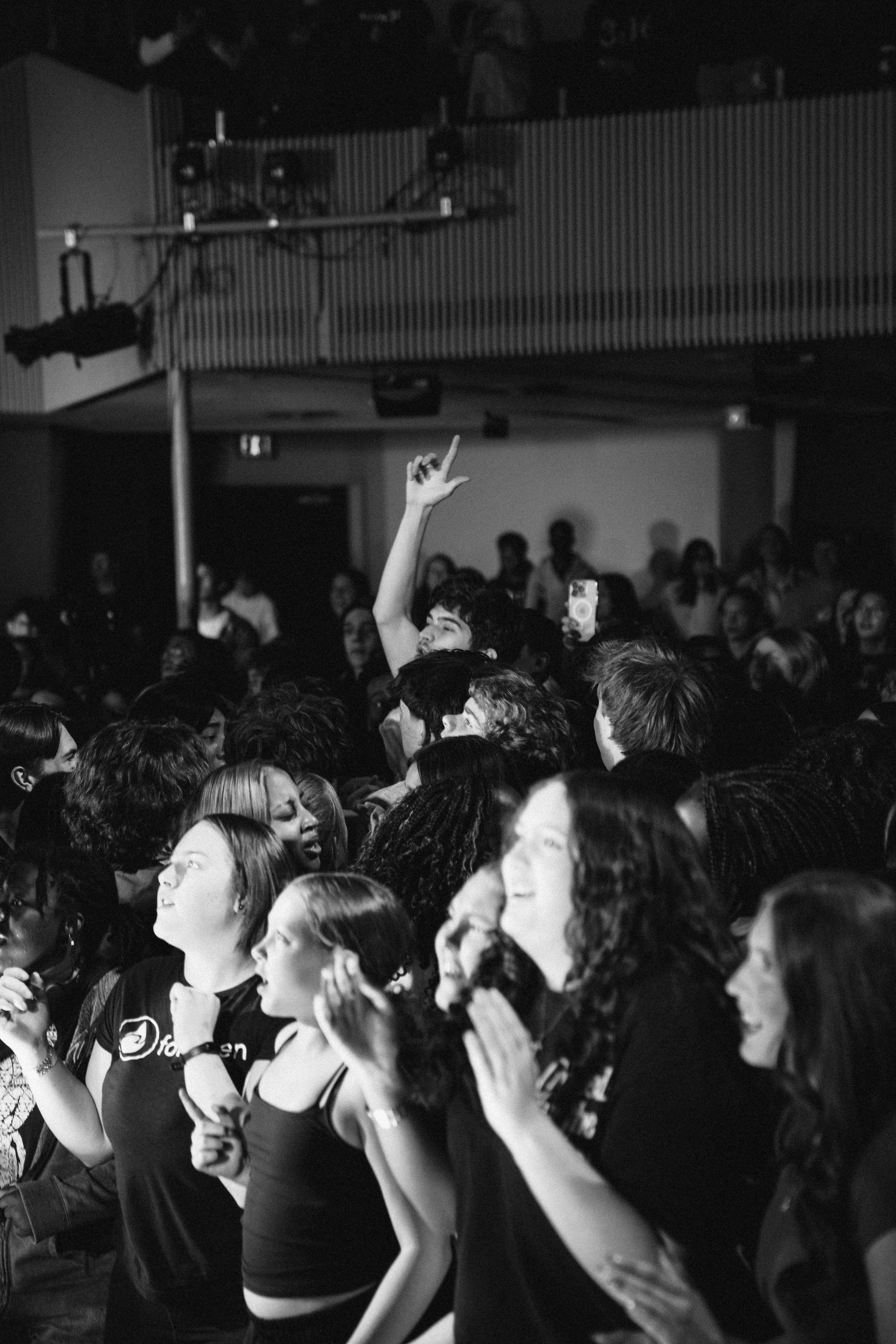 A crowd of diverse young people standing together in a public space, some singing or clapping, with a woman in the foreground raising one hand, holding a phone, and looking determined.