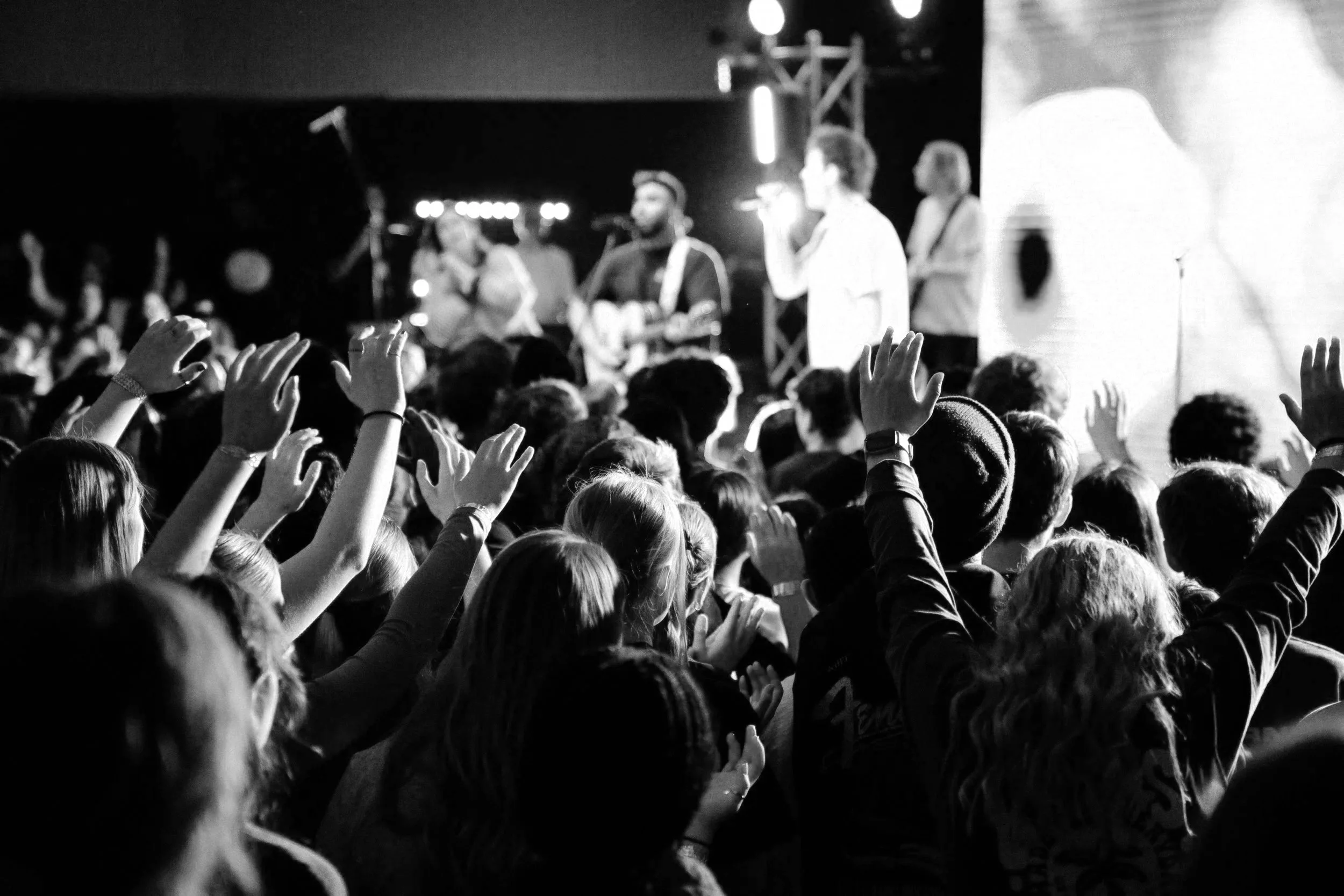 A black and white photo of a concert with a crowd of people raising their hands and a band performing on stage.