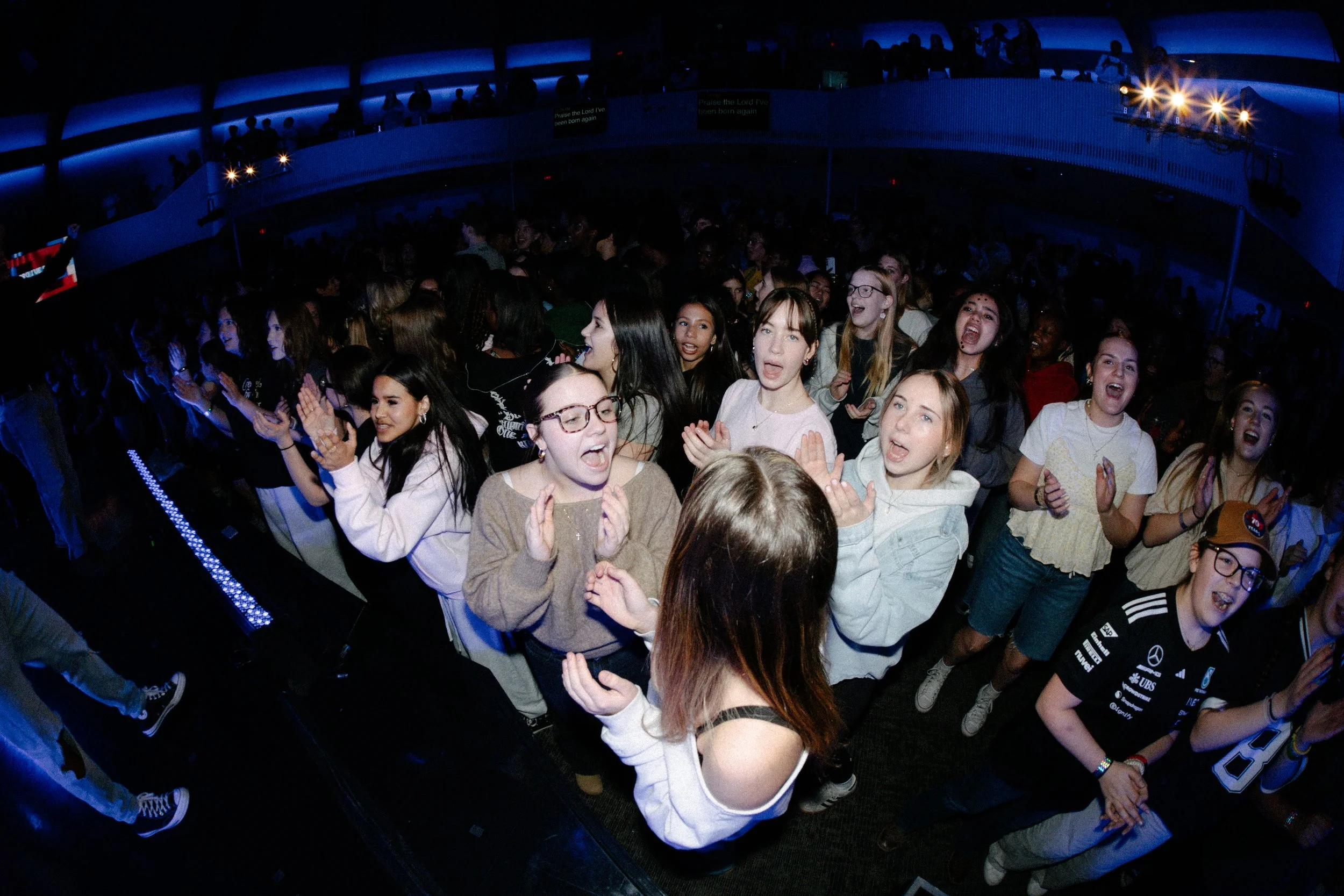 A large crowd of young people enjoying a concert or event in a dimly lit indoor venue with purple-blue lighting.