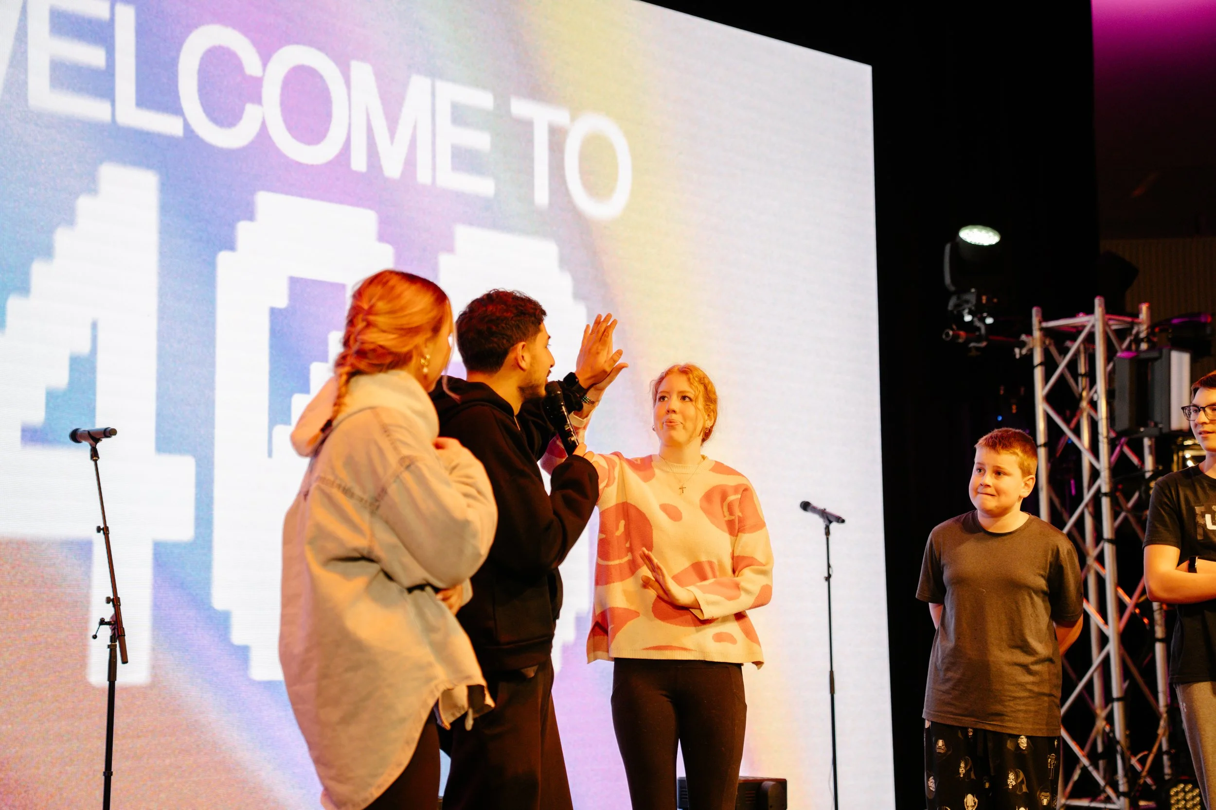 People on stage during an event with a large screen in the background displaying the words 'WELCOME TO' and a pixelated cityscape image. Five individuals, including children and adults, are engaged in conversation or presentation, with microphones an