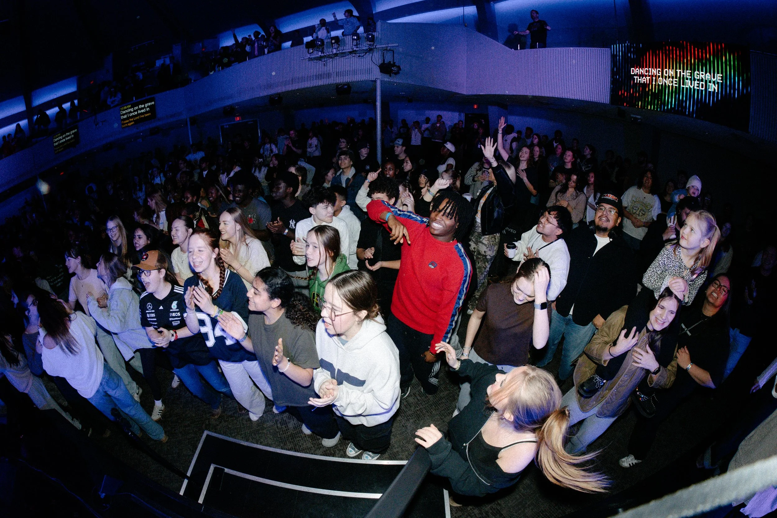 A large crowd of people at a concert or event, with some on the floor and others on a balcony, dancing and enjoying the music. There is a sign on the balcony that reads 'DANCING ON THE GRAVE THAT I ONCE LIVED IN'.