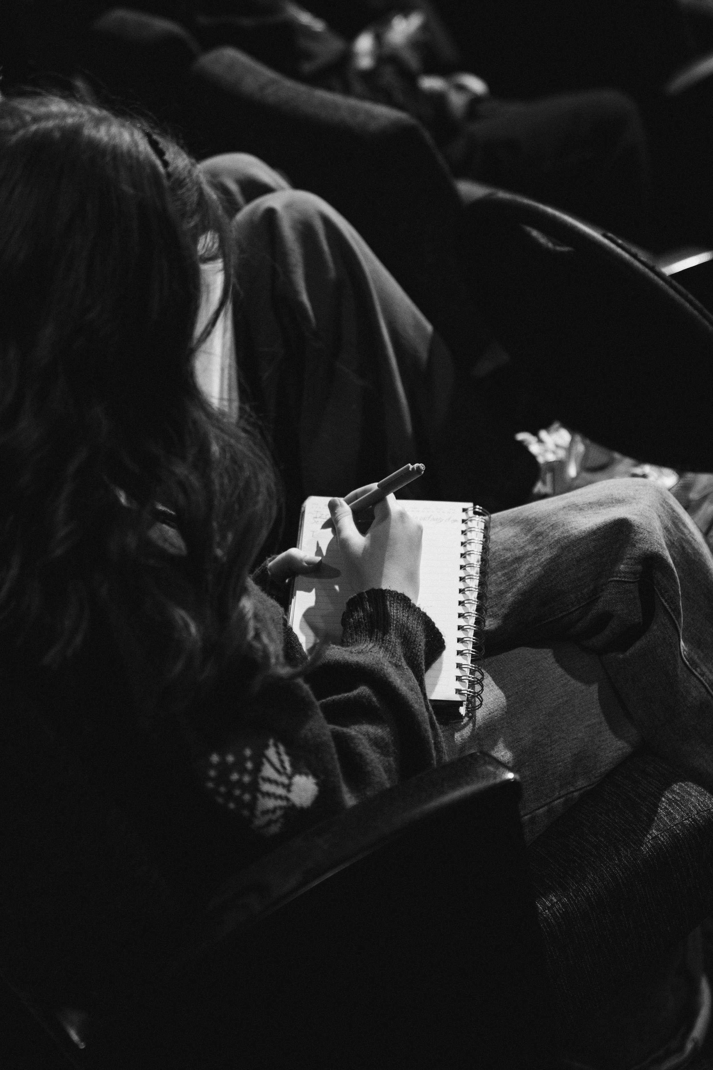 A person with long hair is sitting and writing or drawing in a spiral notebook with a pen, in a dark setting. The person is wearing a sweater with a Christmas pattern on the sleeve.