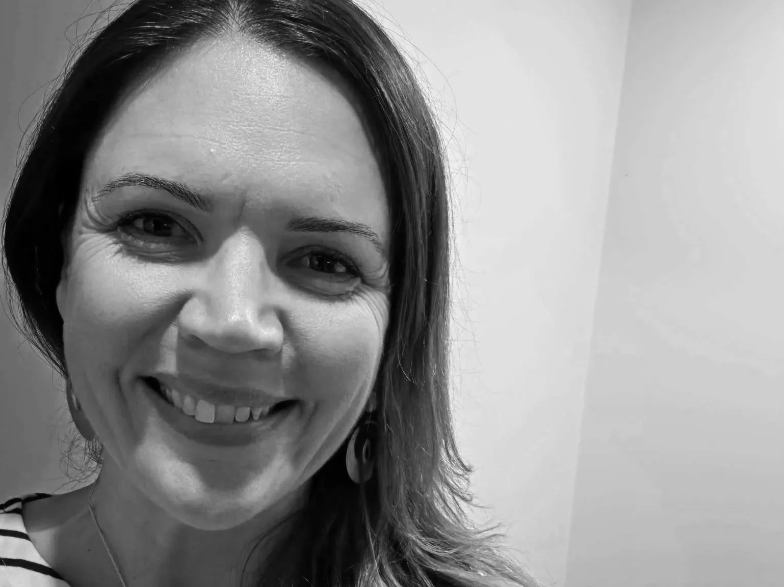 A close-up black and white photograph of a woman smiling, with shoulder-length hair, wearing earrings, and a striped top, standing against a plain background.