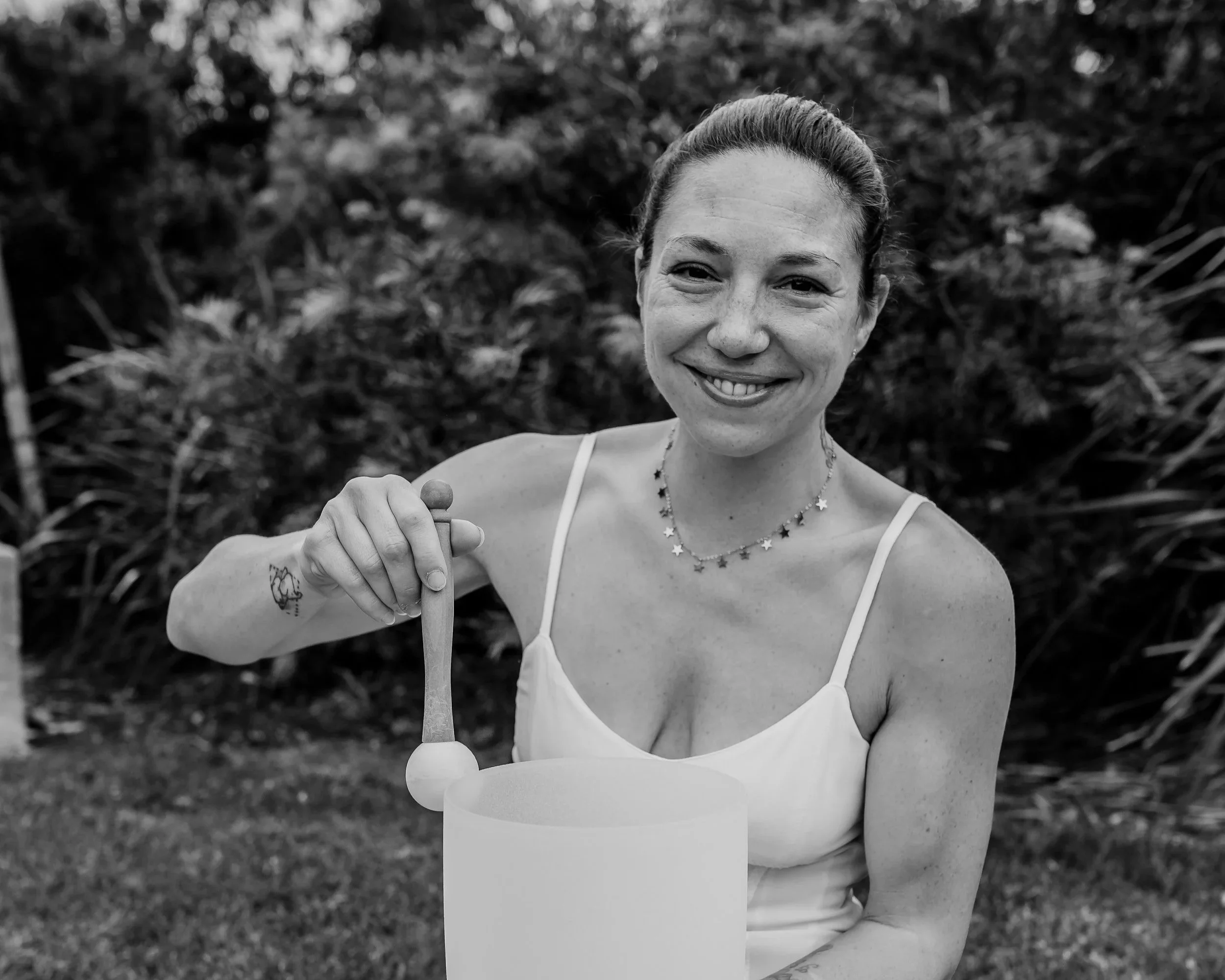 A smiling woman with a ponytail wearing a white tank top and star-shaped necklace, holding a stick of cotton candy outdoors, with a background of foliage.
