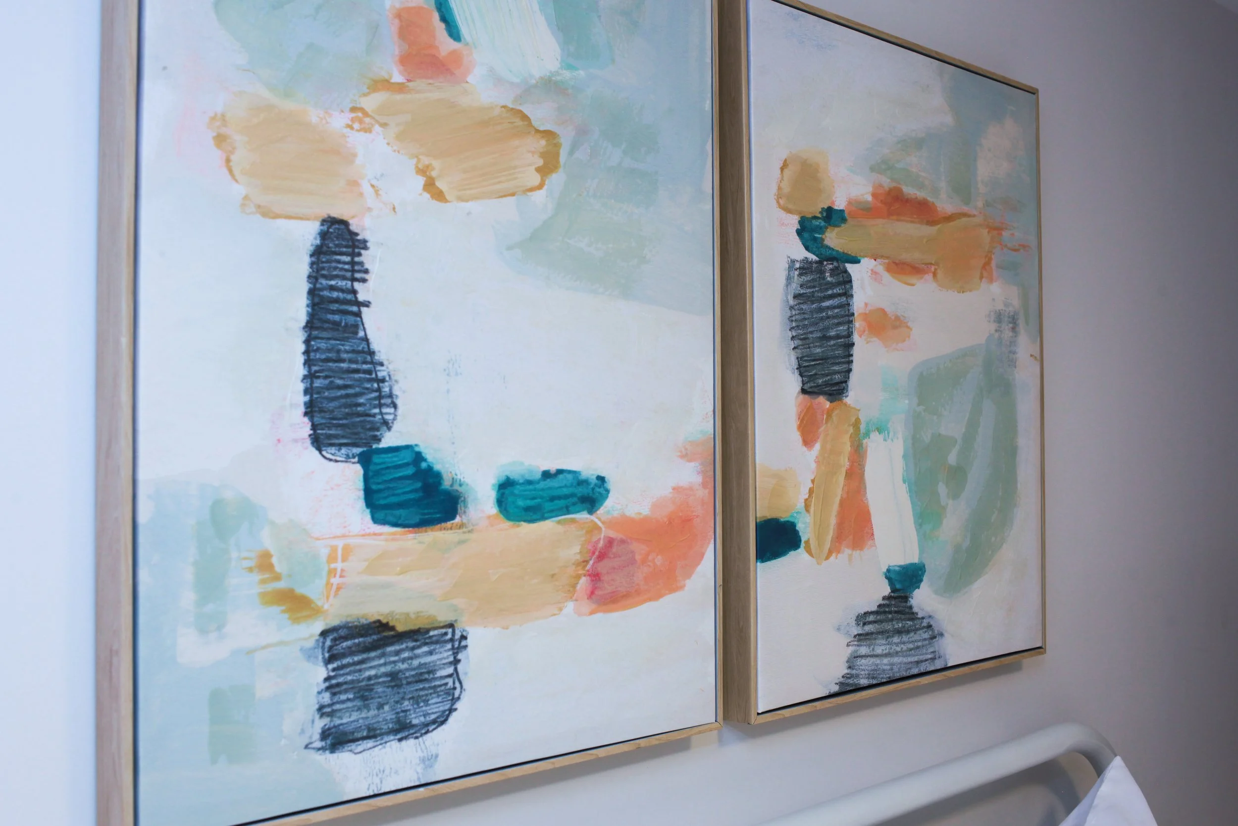 Two abstract paintings with pastel colors, black and teal brushstrokes, framed and hung side by side on a wall.