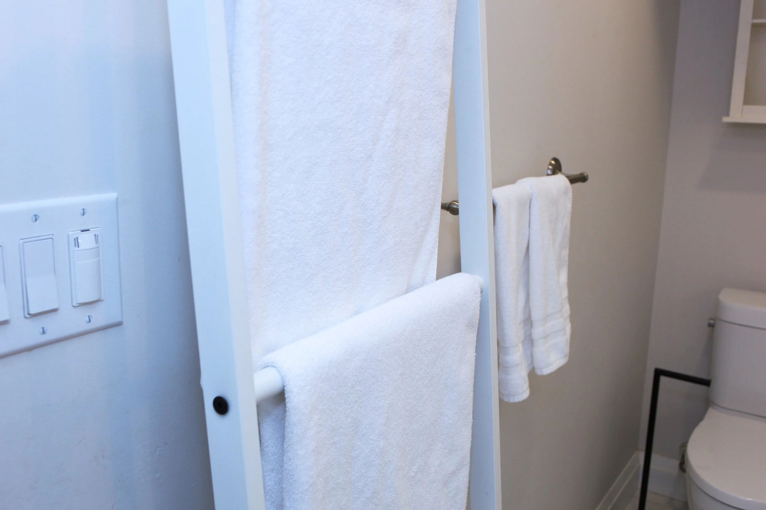 A close-up of a bathroom with white towels hanging on a towel rack, a light switch on the wall, and a toilet in the background.