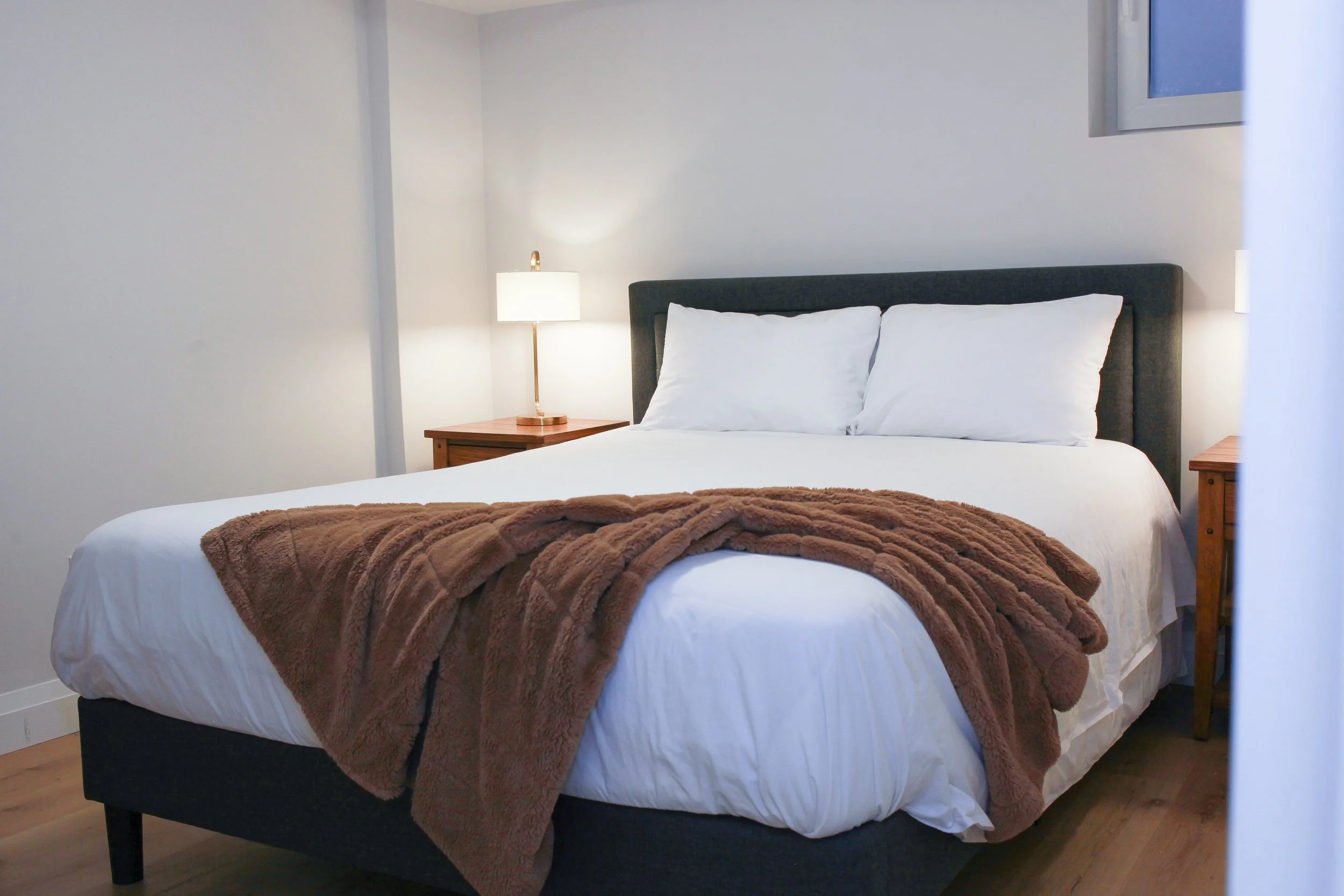 A neatly made bed with white sheets, two pillows, and a brown blanket at the foot of the bed. There are two wooden nightstands with a lamp on the left side of the bed and an open window on the upper right corner of the wall.