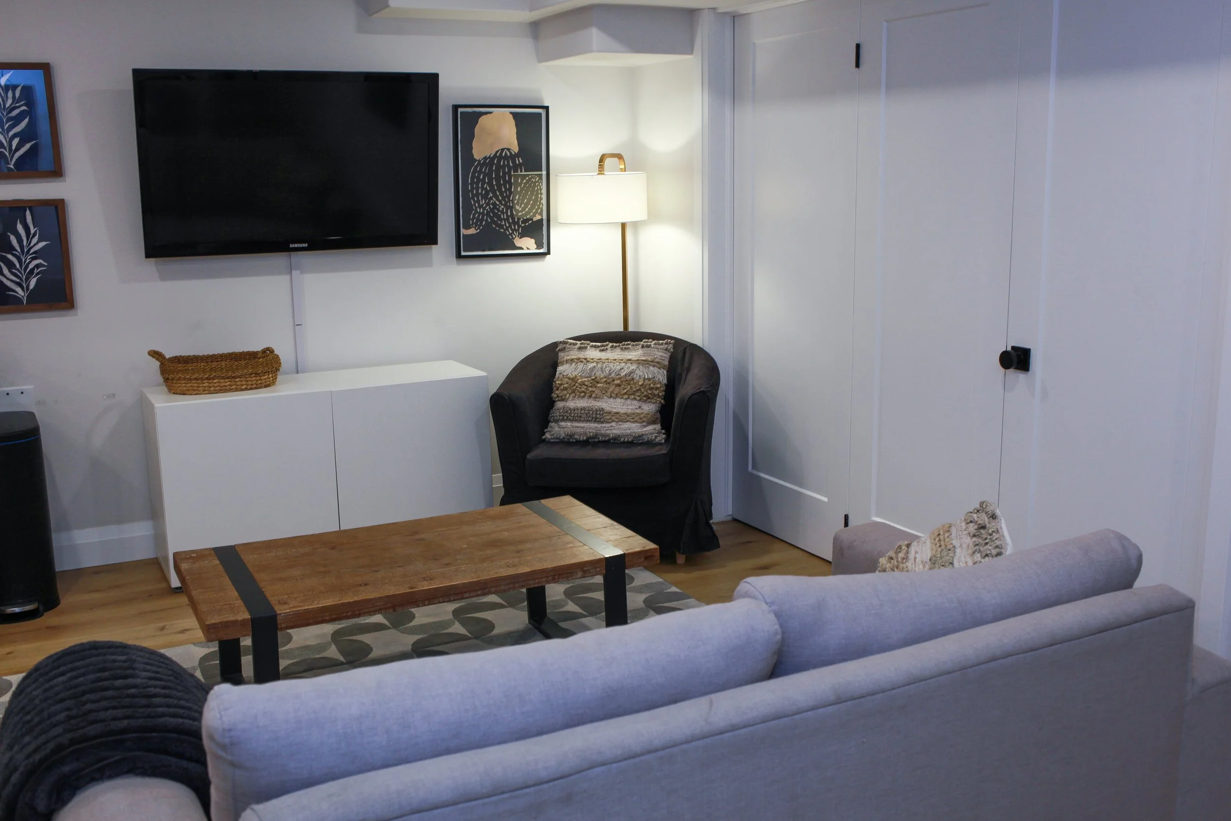 A living room with a wall-mounted television, a white cabinet, framed wall art, a black armchair with a patterned pillow, a wooden coffee table, a gray sofa with a decorative pillow, a folding door, and a floor lamp.