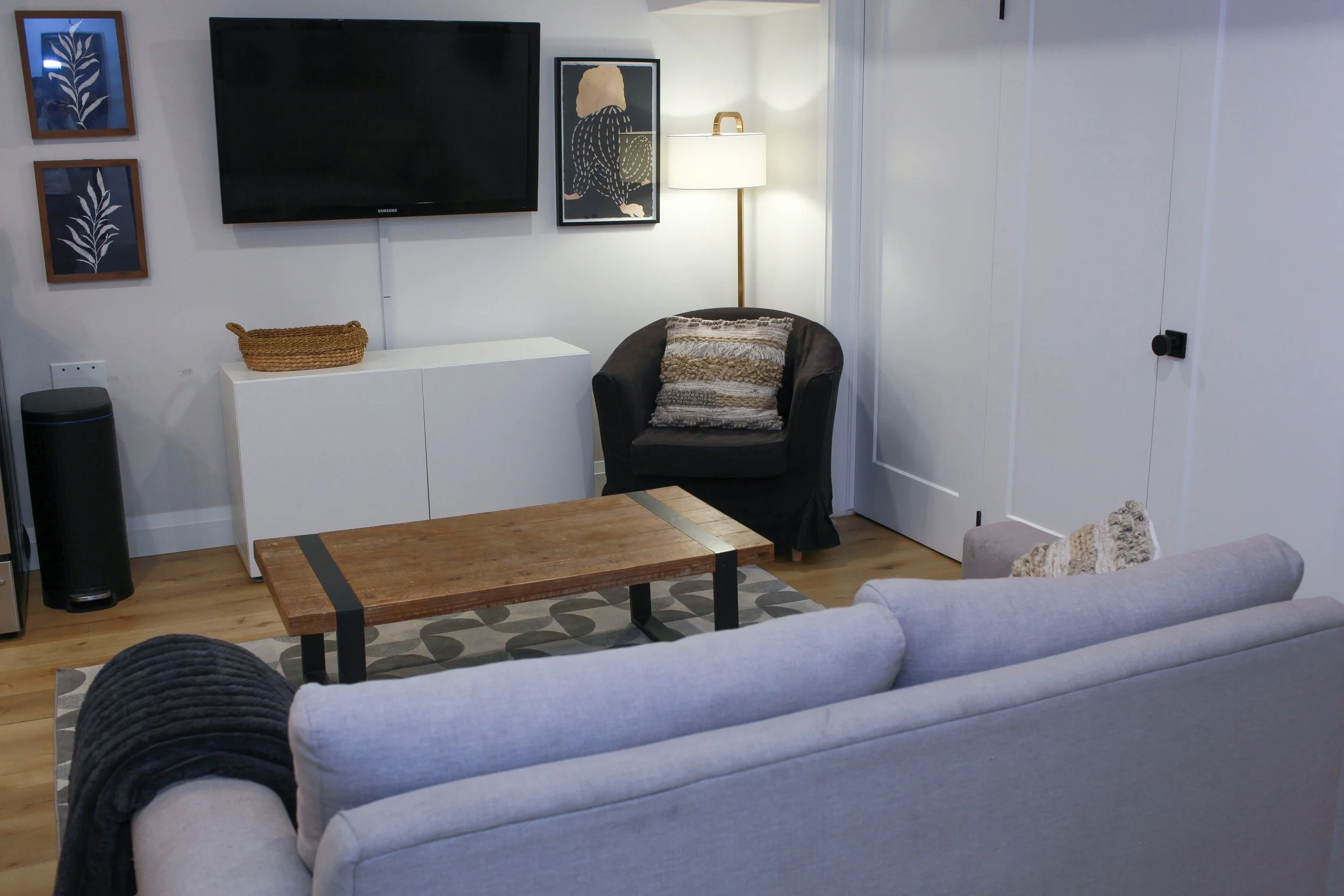 Living room with a beige sofa, coffee table, black armchair with a decorative pillow, white media console, wall-mounted TV, framed artwork, and a floor lamp.