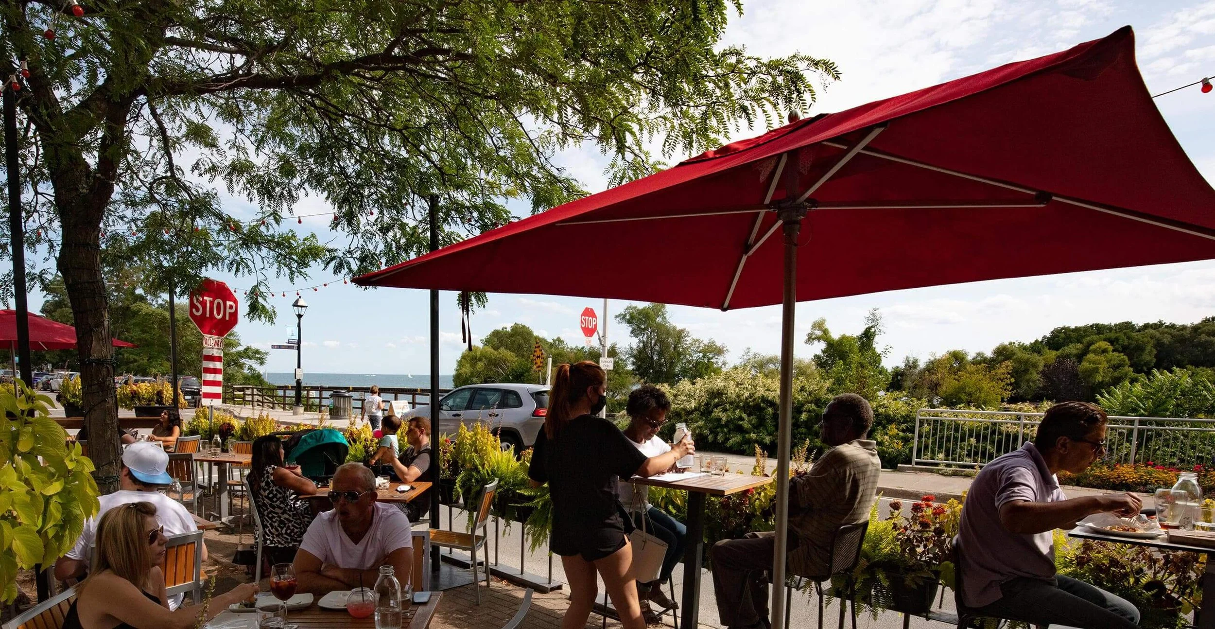 People dining at outdoor patio of a restaurant with red umbrellas, trees, and a view of the water in the background on a sunny day.