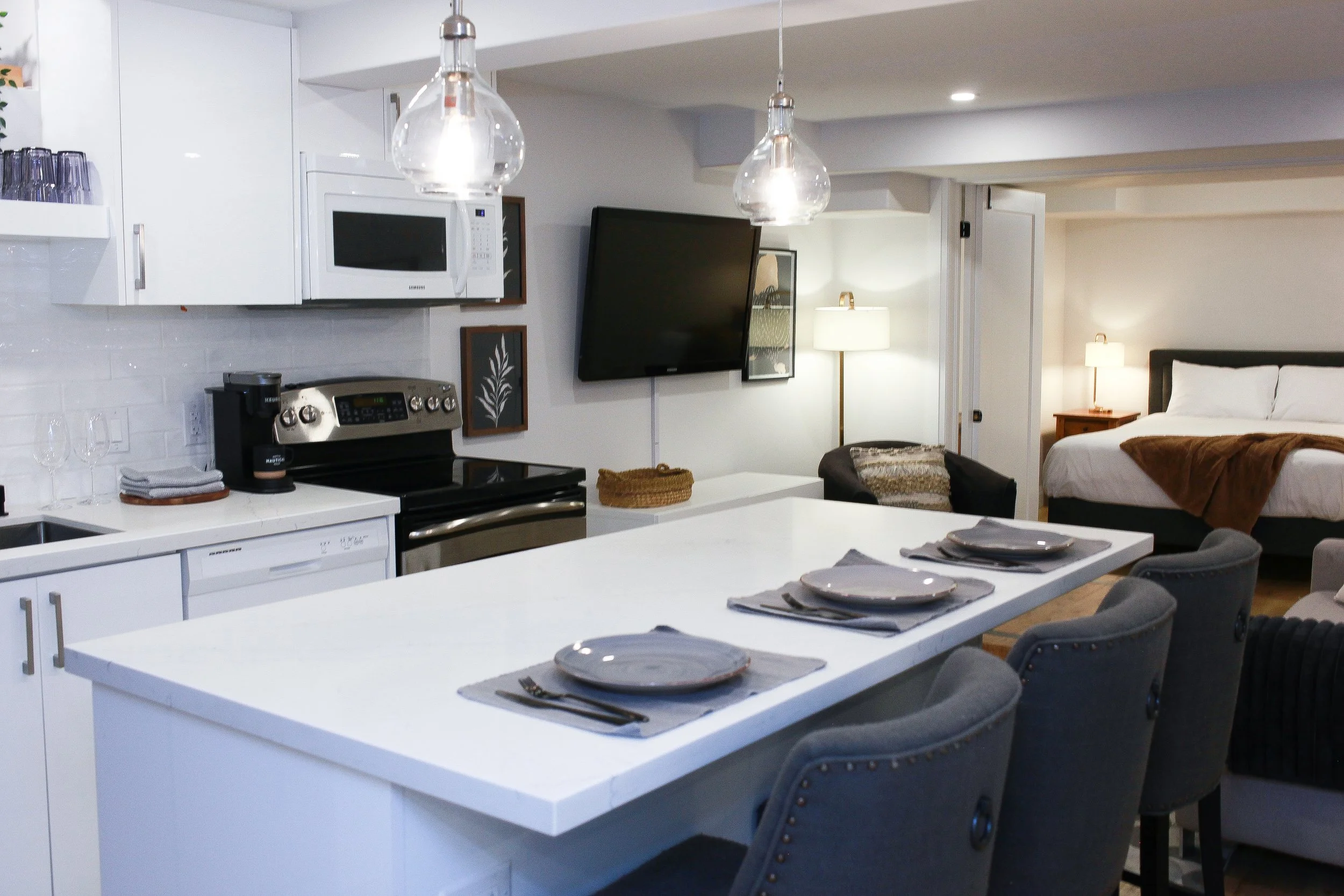 Modern kitchen and living area with white cabinets, a stove, microwave, and coffee maker. A dining table with place settings and chairs. A bedroom with a bed, nightstand, and lamps is visible in the background.
