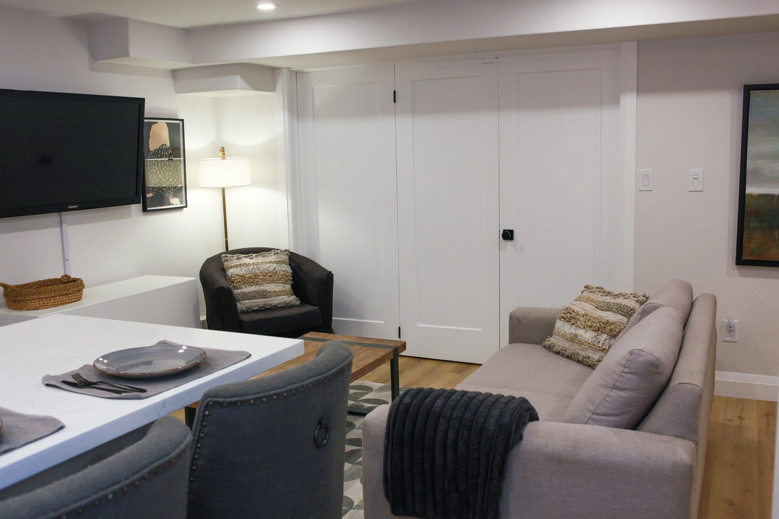 Living room with beige sofa, black armchair, wall-mounted TV, floor lamp, artwork, and a closed white closet door.