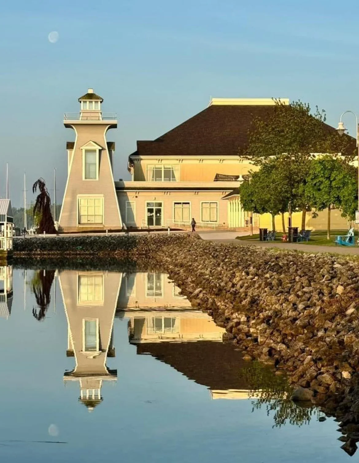 A building with a lighthouse-style tower near water with a reflection, trees, benches, and a person walking nearby, under a partly cloudy sky with the moon visible.