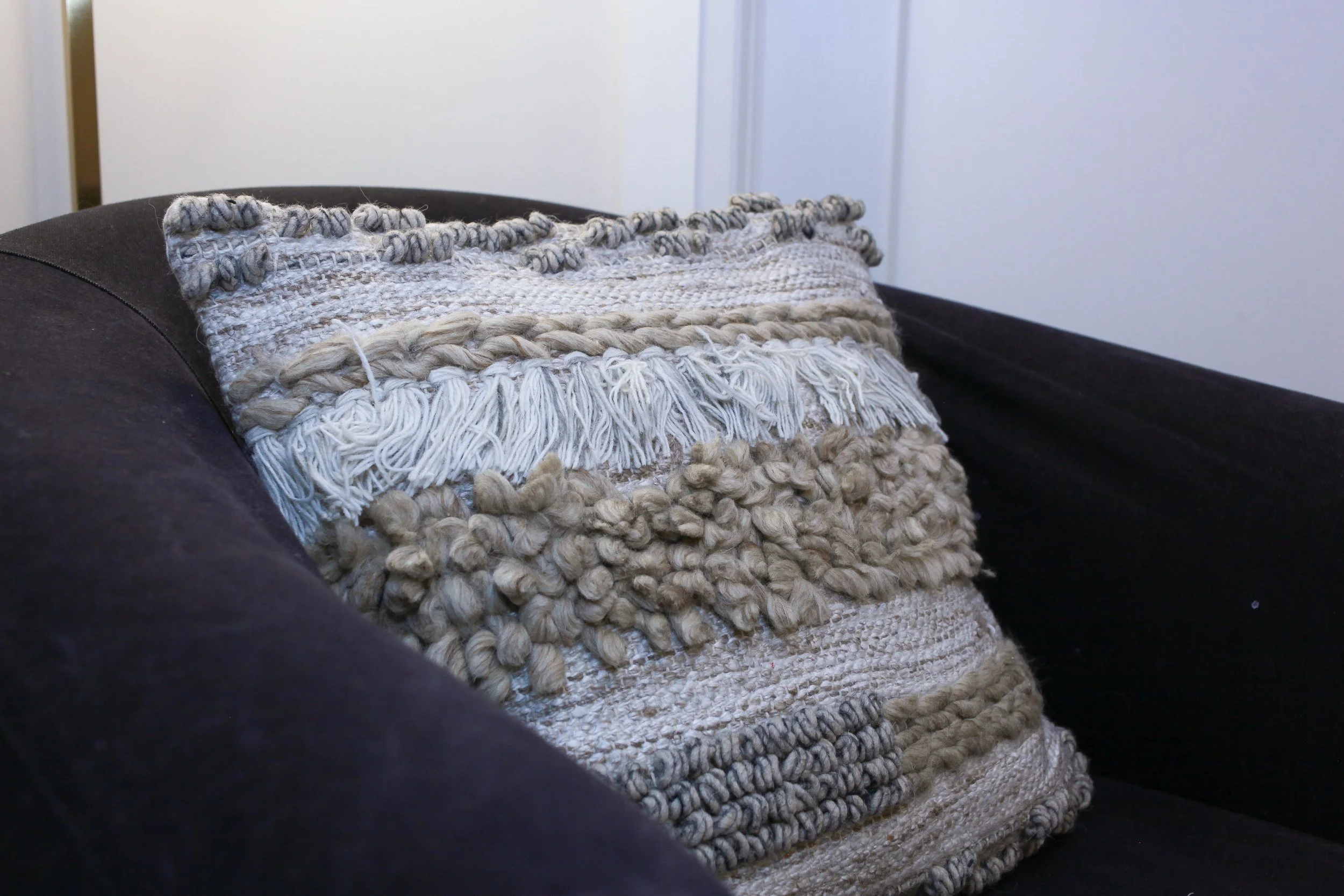 Close-up of a decorative pillow with textured, woven, and knotted fabric in neutral tones on a black couch.