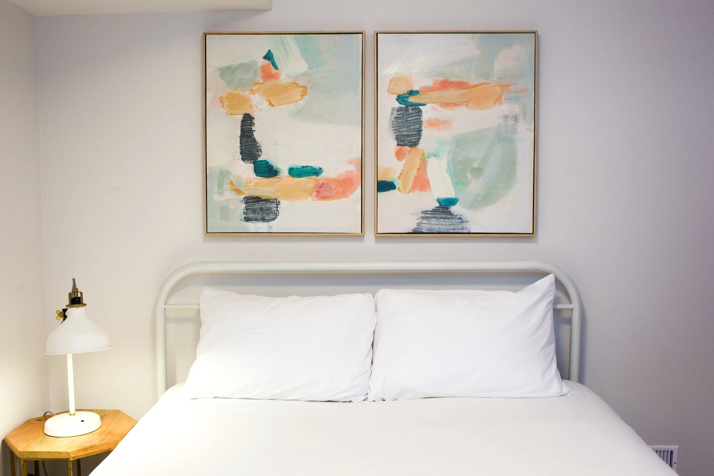 A minimalist bedroom with a white bed, white pillows, a white metal headboard, a wooden bedside table, a white table lamp, and two abstract paintings above the bed with pastel colors.