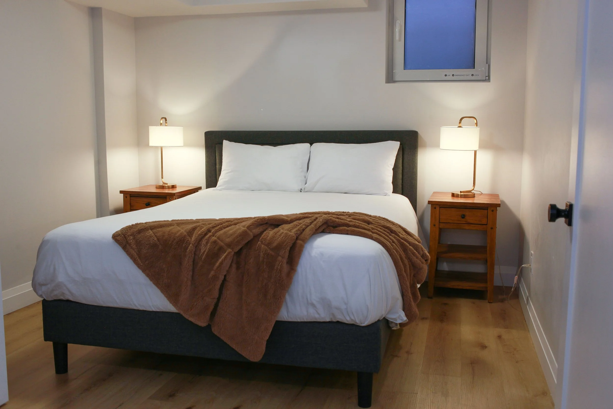 A neatly made bed with white sheets and two white pillows, topped with a plush brown blanket, in a bedroom with two wooden nightstands and lamps, and a wall-mounted flat-screen TV.