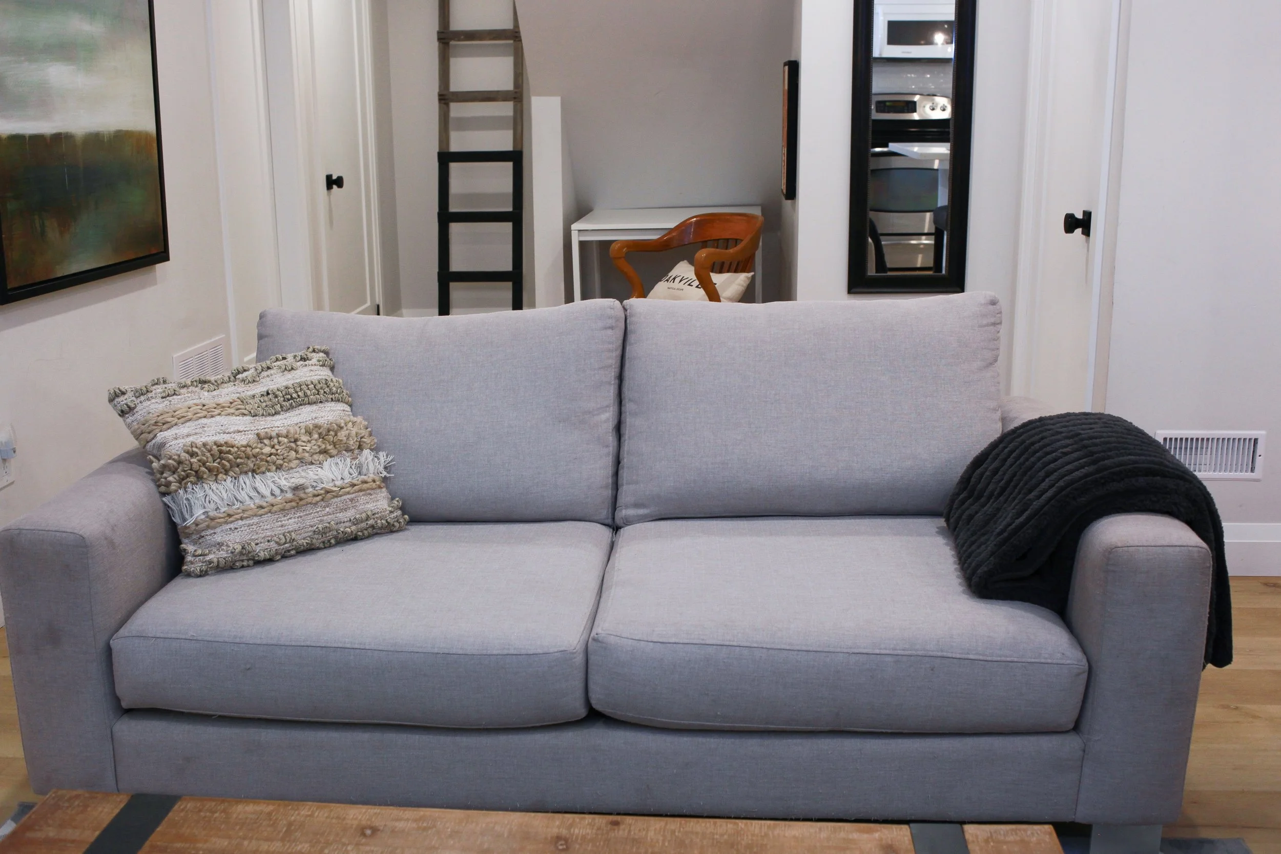A light gray fabric sofa with two cushions, one textured with stripes and fringes on the left, and a black blanket draped over the right armrest, in a cozy living room.