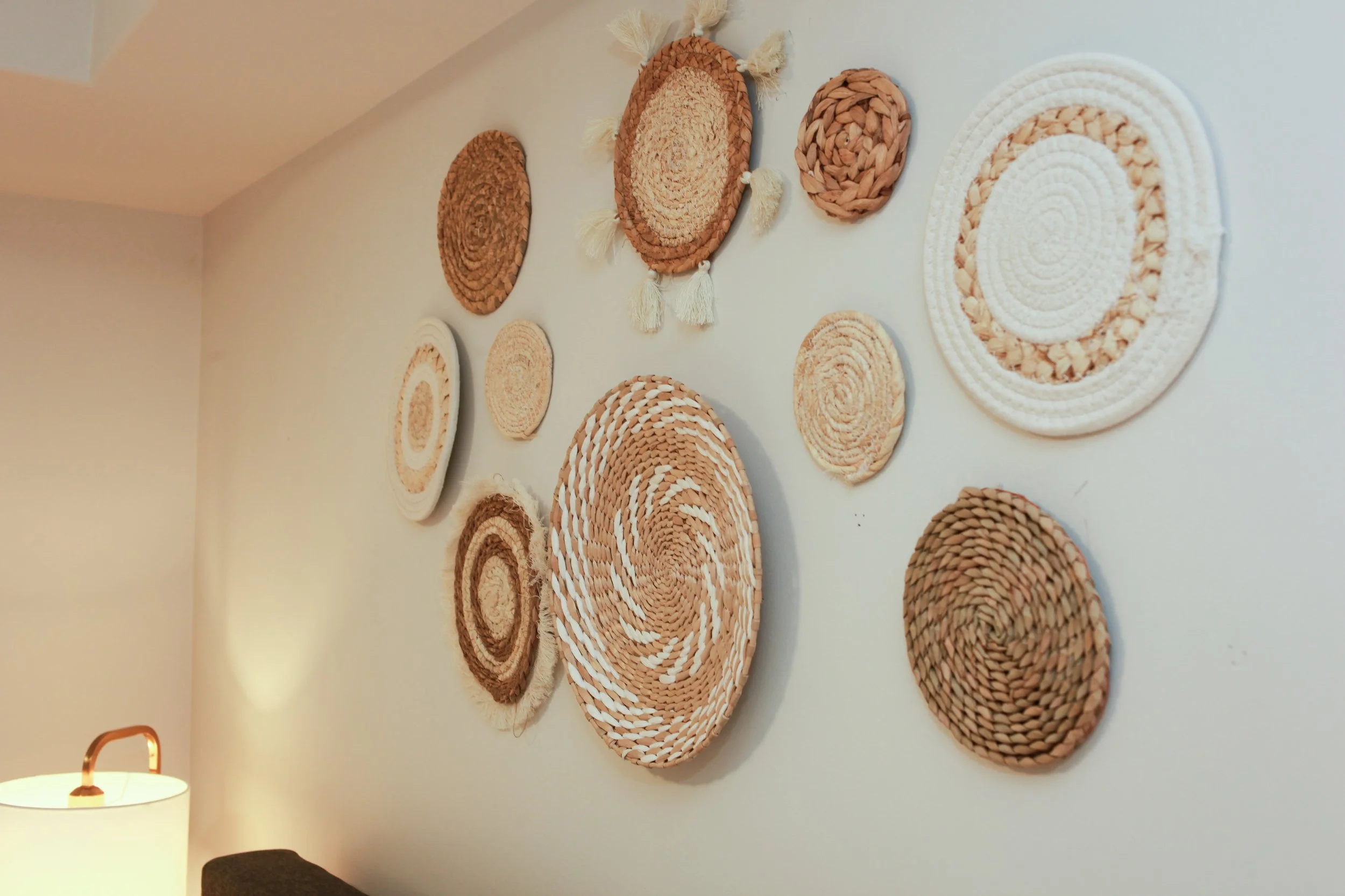 Decorative woven wall art pieces in various shapes and sizes arranged on a white wall, with a lamp and a dark-colored furniture piece in the lower left corner.