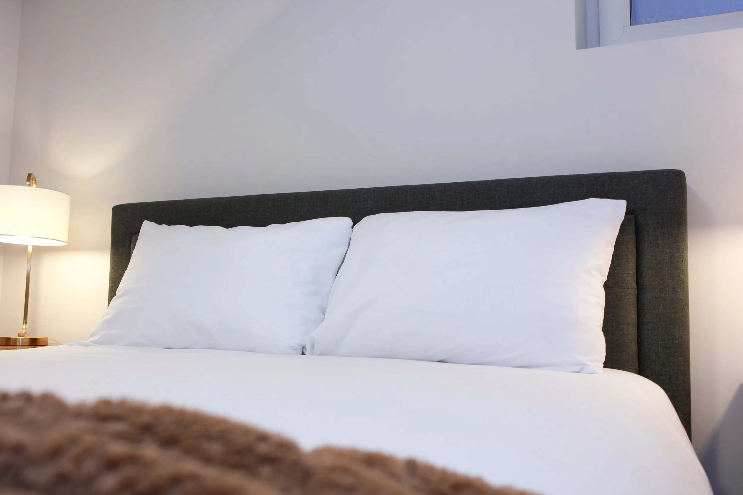 A neatly made bed with white pillows and sheets, a dark gray headboard, a cream-colored lamp on the bedside table, and a small window in the corner of the wall.