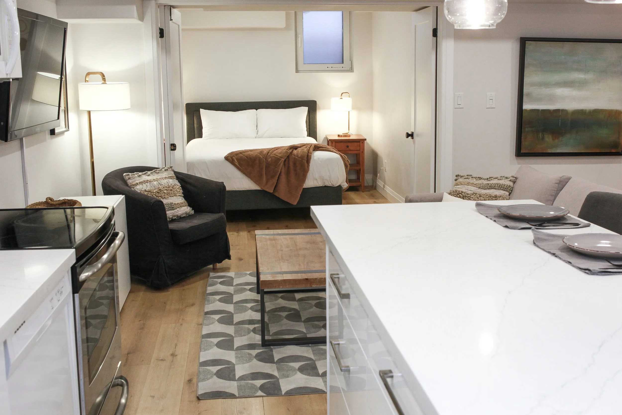 Open-concept studio apartment with kitchen, living area, and bedroom. The kitchen has white cabinets, a white marble countertop, and a stove. The living area features a black armchair with a patterned pillow, a coffee table, and a patterned rug. The 