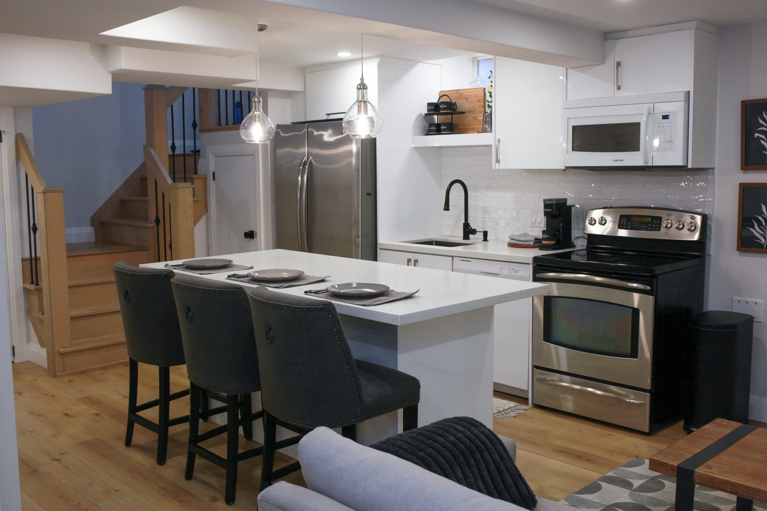 Modern kitchen with white cabinets, stainless steel appliances, black faucet, white countertop island with three black chairs, and black pendant lights.