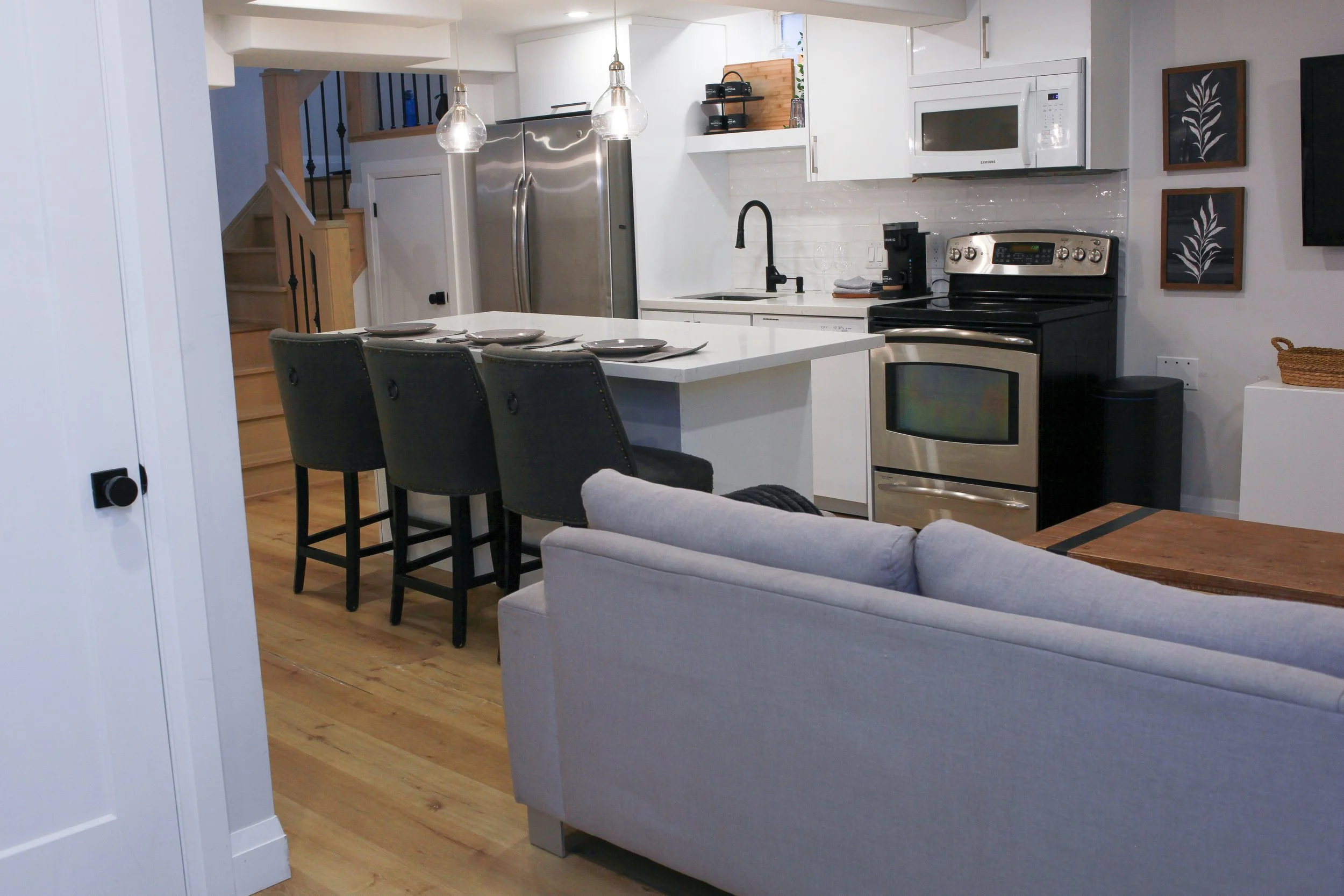 Modern open-concept kitchen with white cabinets, stainless steel refrigerator, black stove, microwave, and black bar stools at a white kitchen island with three place settings. A beige sofa is in the foreground, and the kitchen features wooden floori