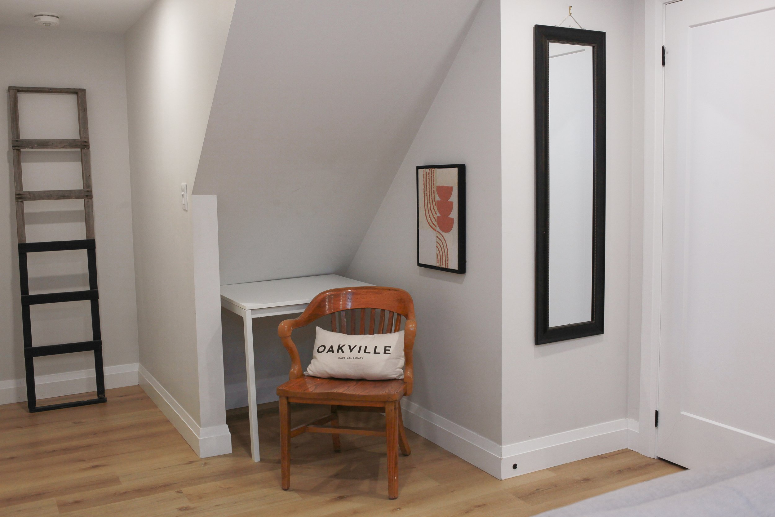 A corner of a room featuring a wooden chair with a pillow that reads "OAKVILLE". There is a small white table behind the chair, two framed pictures on the wall, and a decorative ladder leaning against the wall. The floor is hardwood, and the walls ar