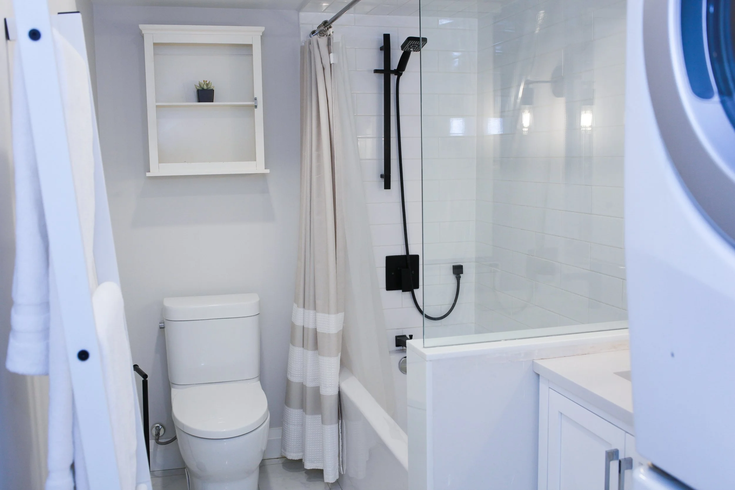 Bathroom with white toilet, shower area with glass partition, beige and white striped shower curtain, black shower fixtures, white wall-mounted cabinet, and washing machine on the right.