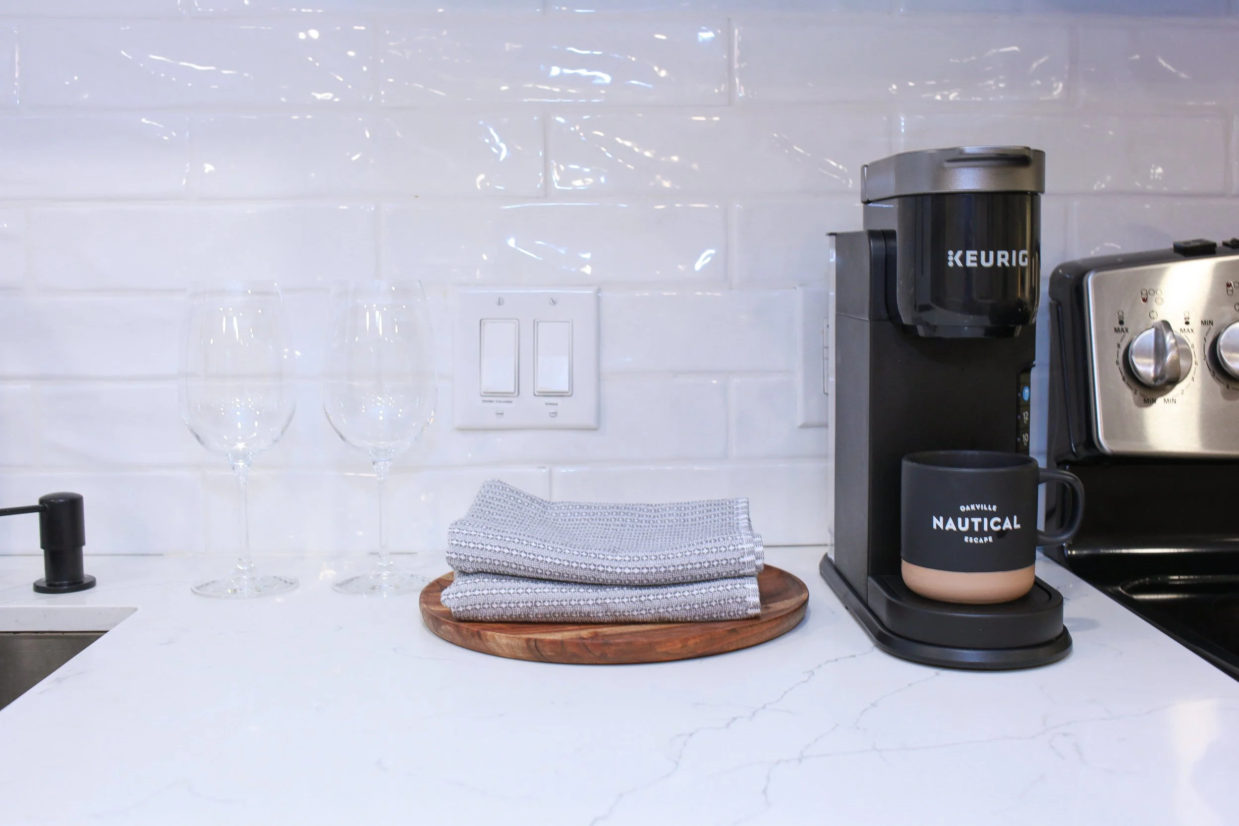 Kitchen counter with two empty wine glasses, a wooden tray with folded gray patterned napkins, a black coffee mug with text 'Oakville Nautical Escape', a Keurig coffee maker, and a stainless steel toaster against a white subway tile backsplash.