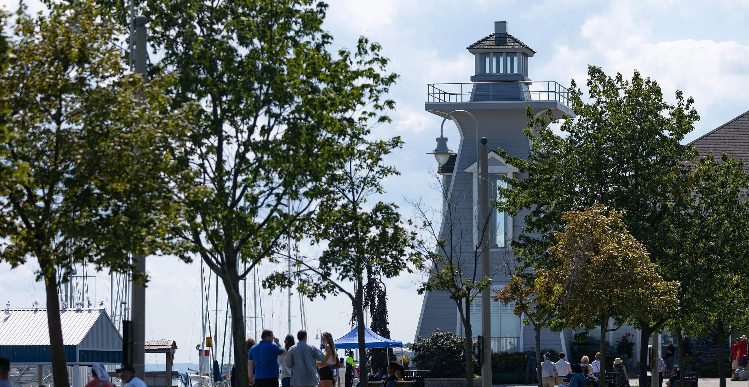 A marina area with sailboats, trees, and a lighthouse-shaped building. People are walking and gathering near the waterfront, some under a blue canopy. The scene is lively with a partly cloudy sky.