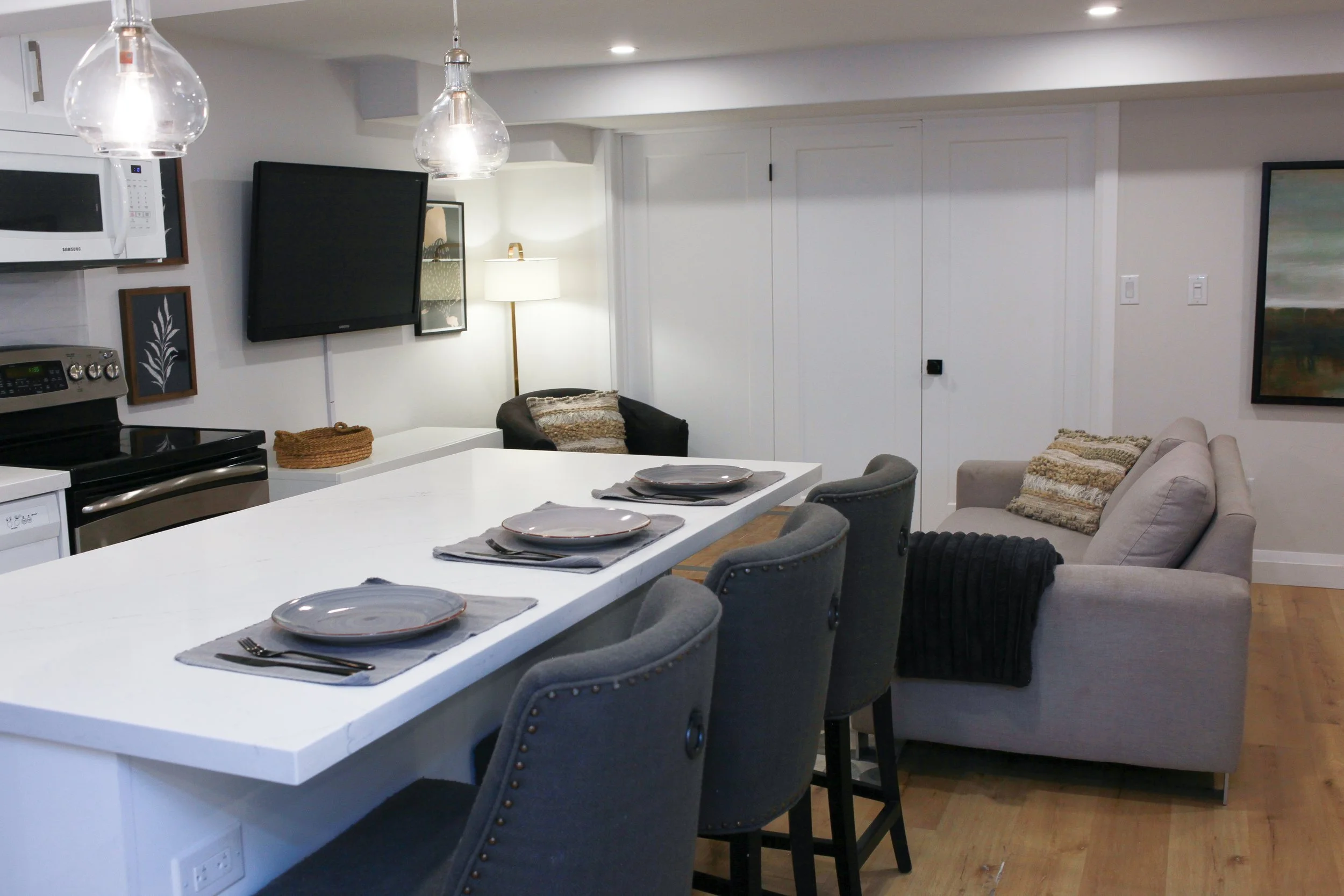 Living and dining room with a kitchen island, chairs, sofa, armchair, wall-mounted TV, artwork, lamp, and hardwood floors.