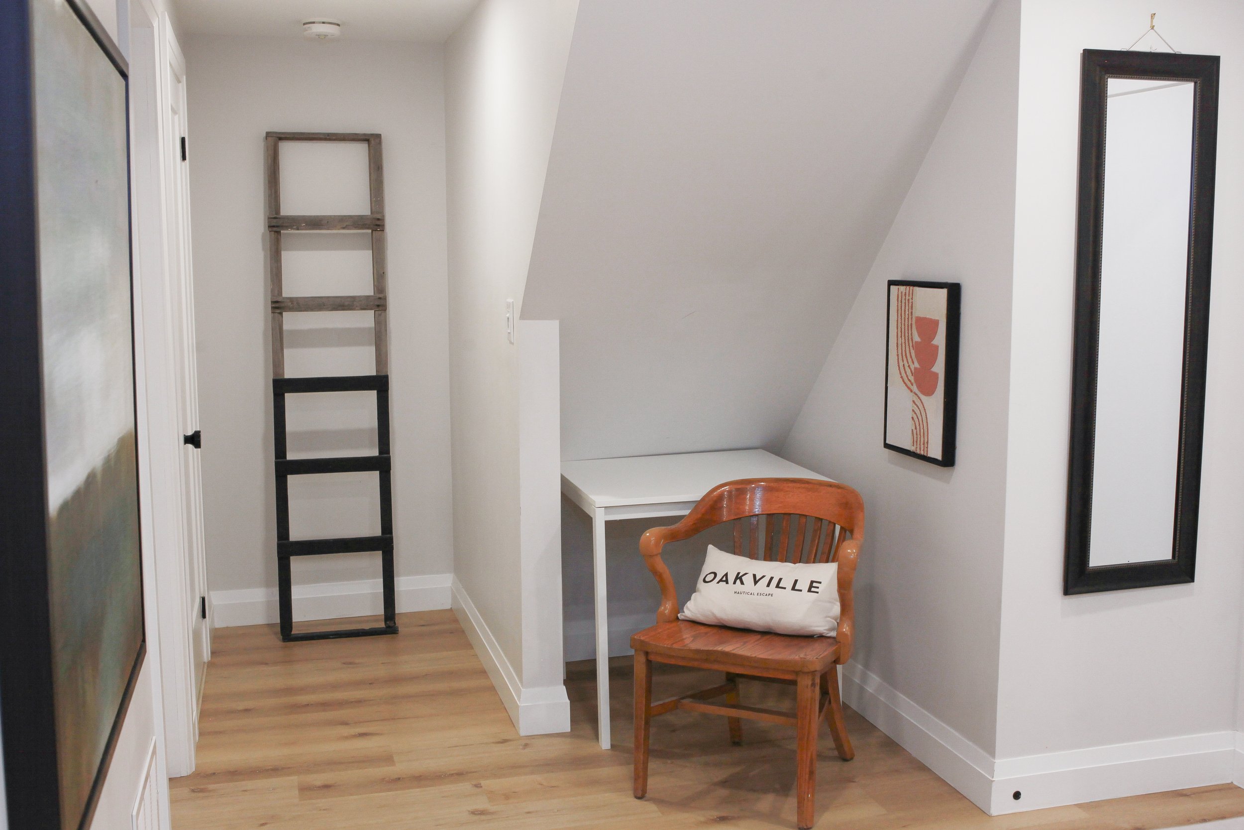A small corner of a room with a wooden chair with a cushion that has 'OAKVILLE' written on it, a white desk, a painted wall with hanging mirrors and art, a wooden ladder, and hardwood flooring.