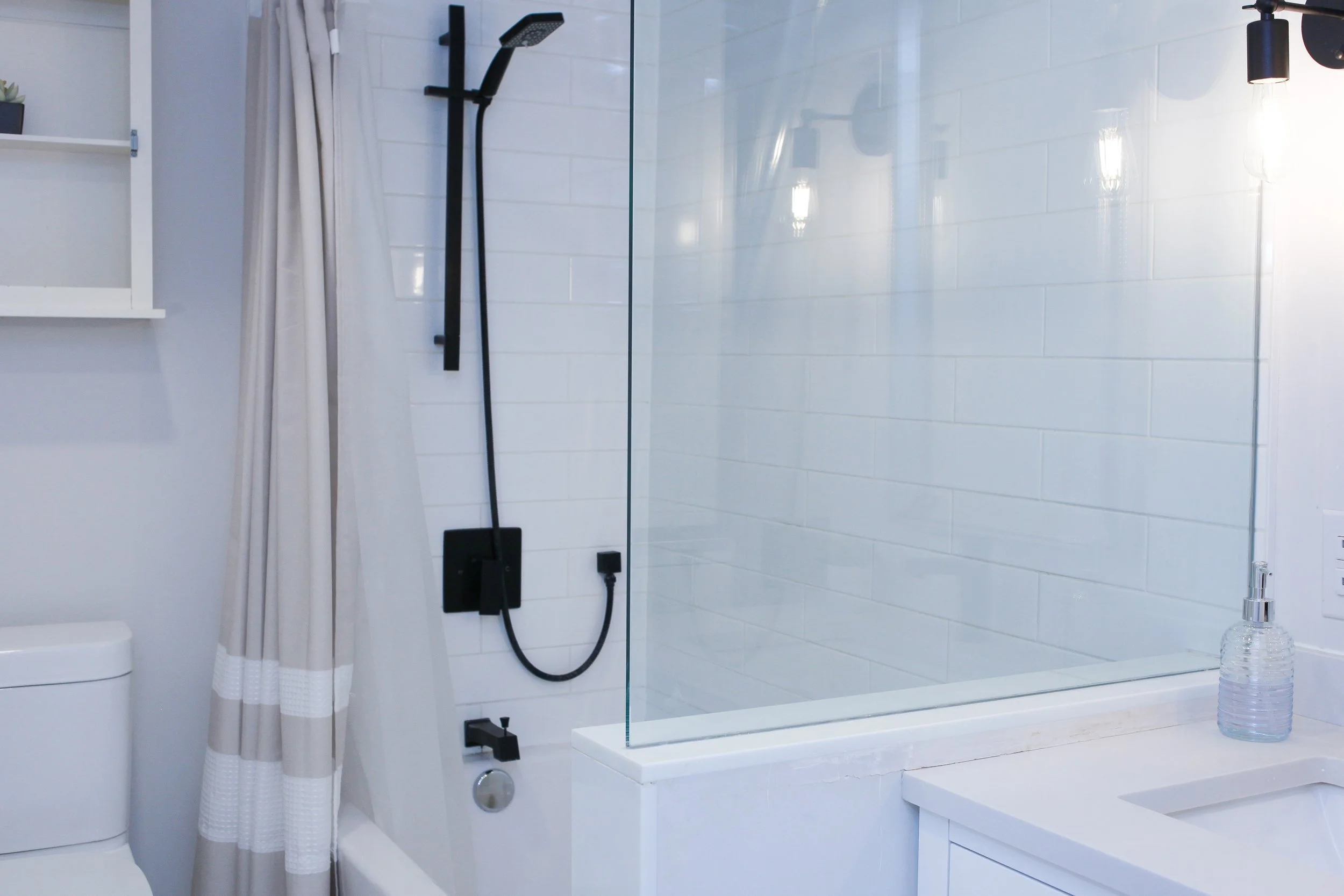 Modern bathroom with white subway tile walls, a glass shower partition, black fixtures, and a beige and white striped shower curtain, with a toilet, a sink, and a clear soap dispenser.
