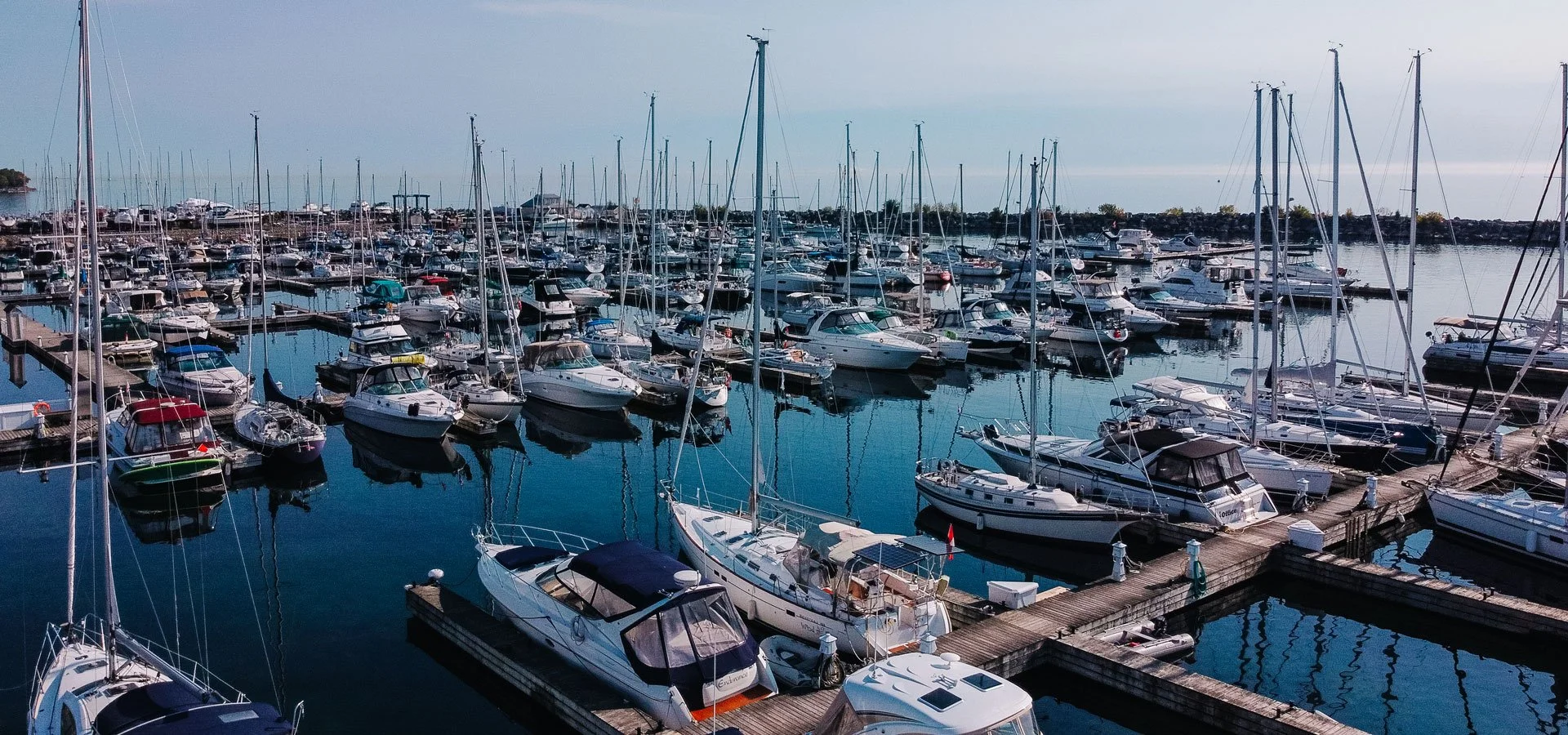 A marina filled with numerous sailboats and motorboats docked at wooden piers, with calm water reflecting the boats and a cloudy sky overhead.