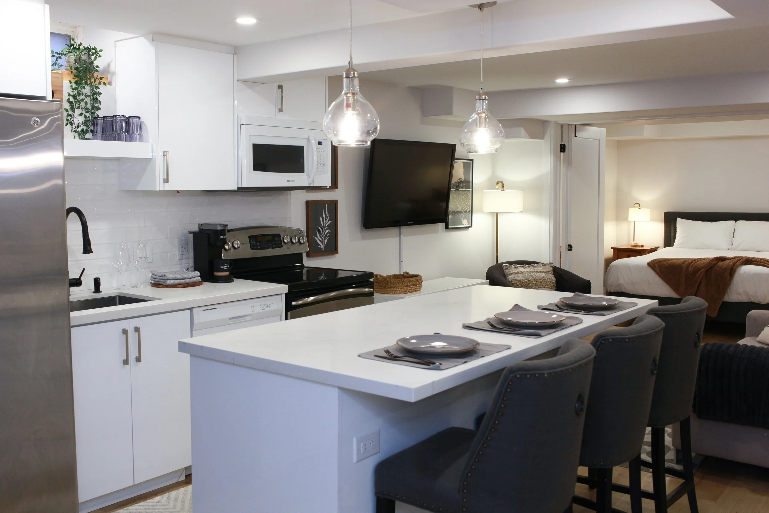 Open-concept kitchen and living area with white cabinets, stainless steel appliances, a wall-mounted TV, a dining table set for four, and a bedroom in the background.