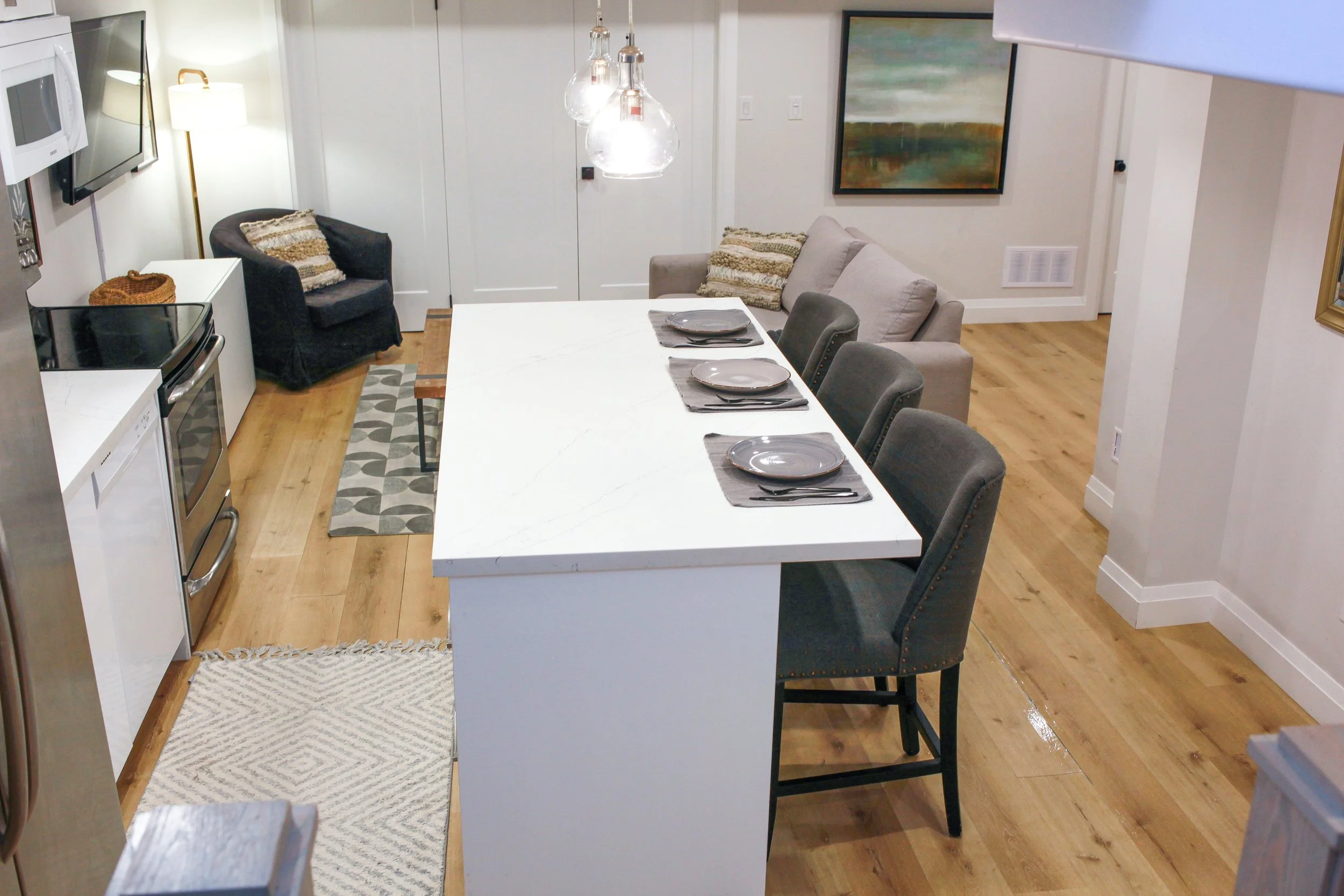 Open-concept living room and kitchen with a white marble kitchen island dining table, four gray upholstered chairs, a beige couch, a black armchair, a painting on the wall, pendant lights, and hardwood flooring.